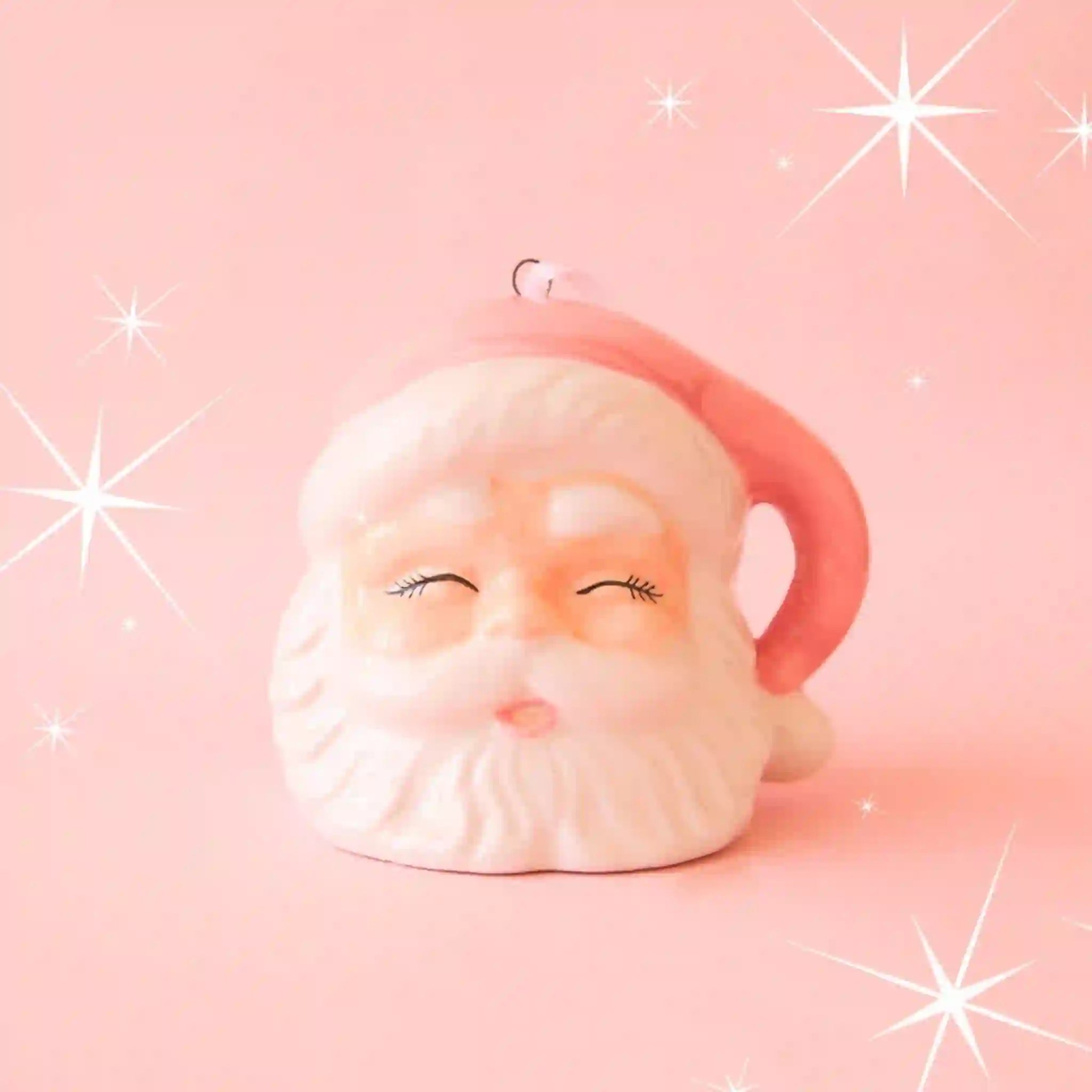 On a pink background is a white and pink Santa head ornament with eyes closed and a pink hat.