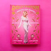 On a bright pink background is a pink book cover with a photo of Dolly Parton and the title that reads, "Dolly Parton Behind The Seams My Life In Rhinestones". 