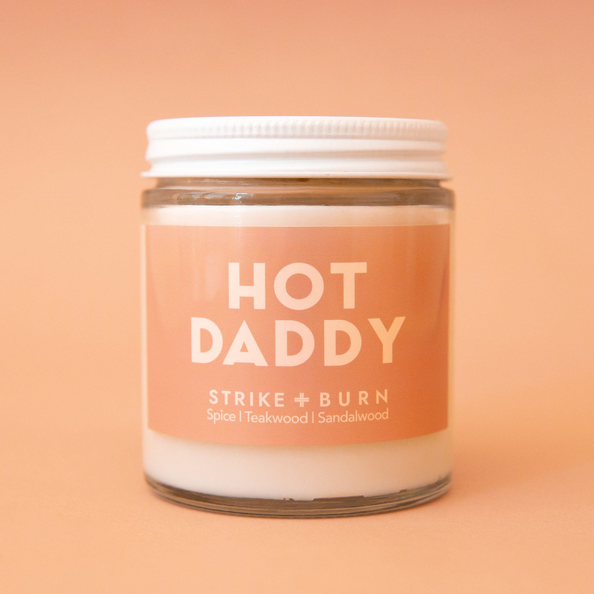 A clear glass jar candle with a light tan label that reads,, "Hot Daddy". 