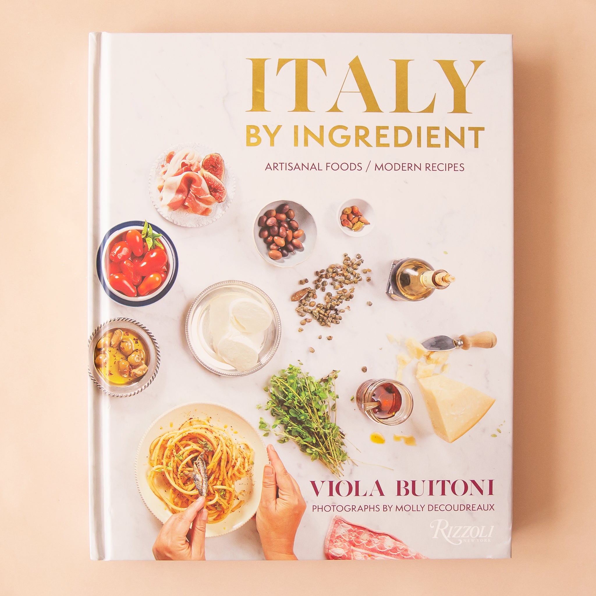 On a peach background is a white book cover with Italian ingredients and the title that reads, &quot;Italy By Ingredient&quot;. 