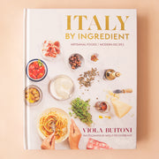 On a peach background is a white book cover with Italian ingredients and the title that reads, "Italy By Ingredient". 