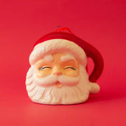 On a red background is a Santa shaped ornament with a red hat on and a closed eye expression. 