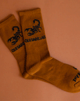 A pair of burnt orange crew socks with a black scorpion graphic and text that reads, "Quesadilla".