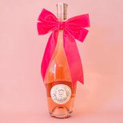 On a peachy background is a wine bottle with a large velvet pink bow