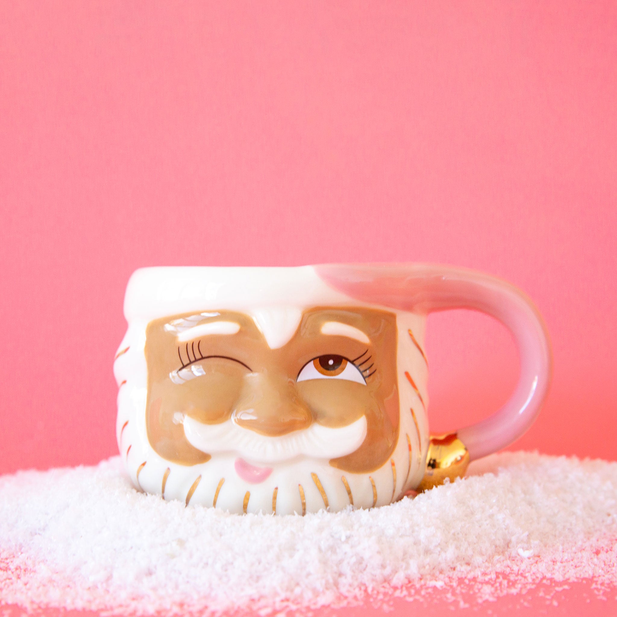 On a pink background is a papa noel face shaped mug with a left eye wink expression and a pink santa hat shaped handle.