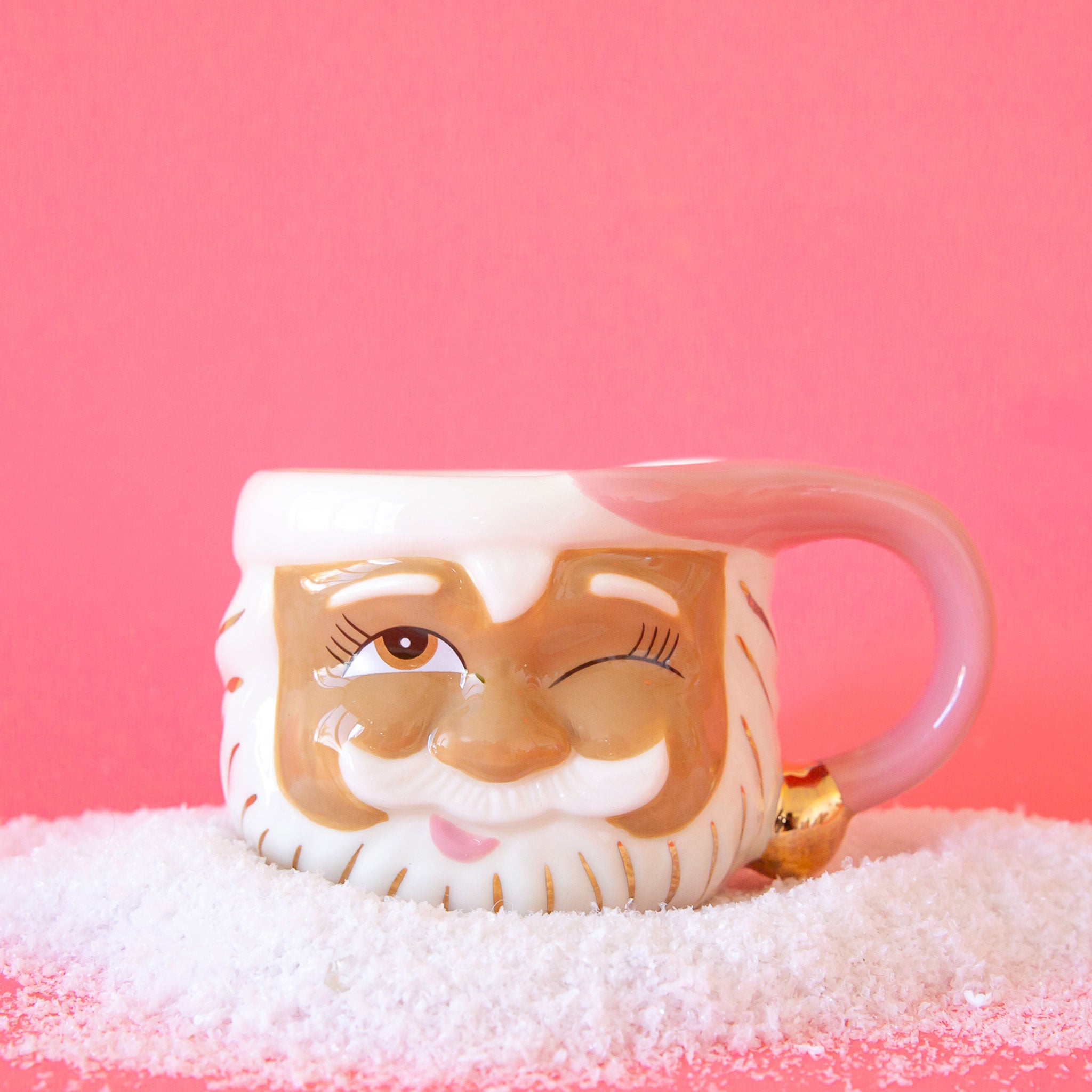 On a pink background is a papa noel face shaped mug with a left eye wink expression and a pink santa hat shaped handle.