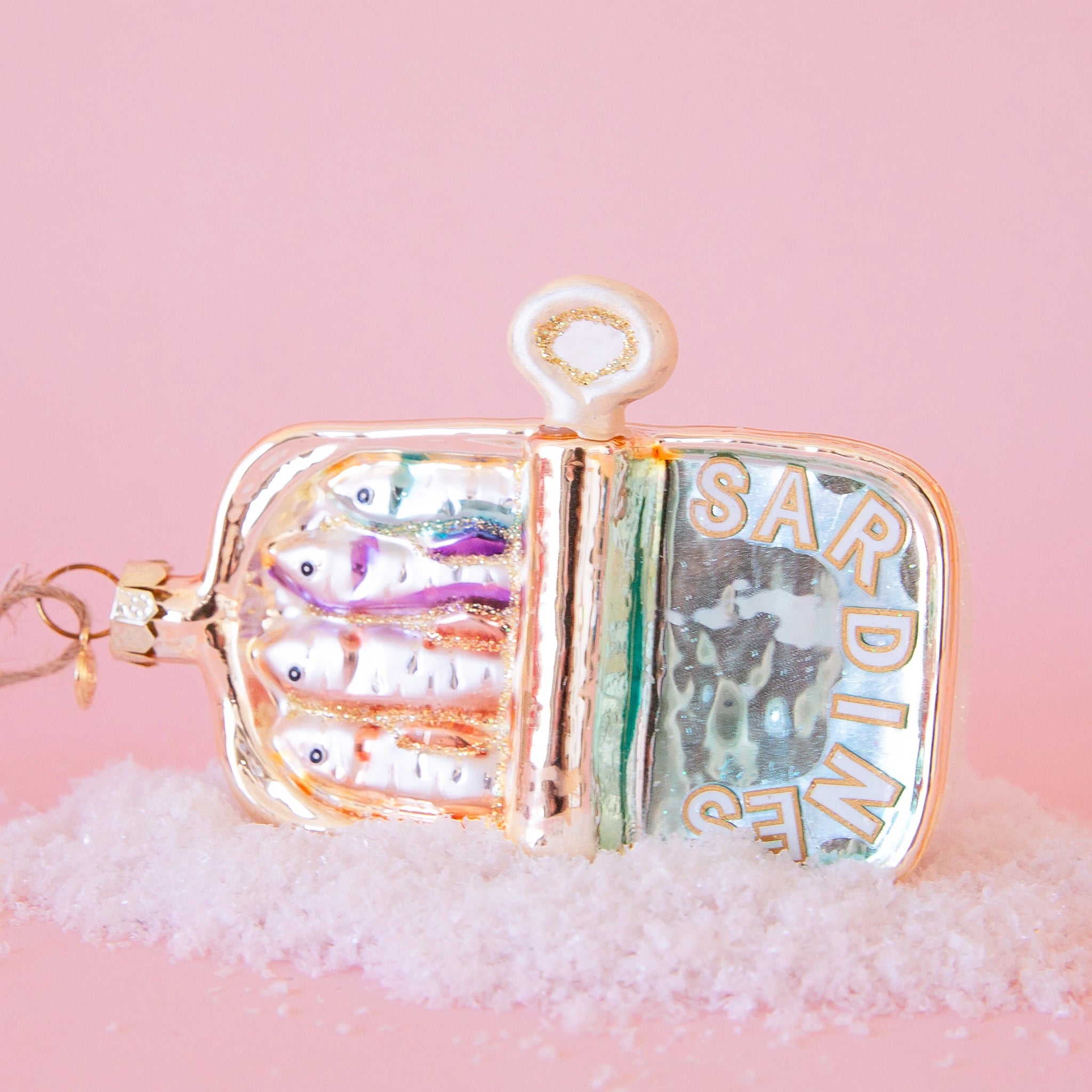 In front of a pink background is a rounded rectangular glass ornament. The ornament is gold metallic. The bottom half is a silvery blue with white text that is outlined in gold. The text reads ‘sardines.’ The bottom half is four heads of sardines. On the right side of the ornament is a white and gold handle. On the top end of the ornament is a gold hook with brown twine.