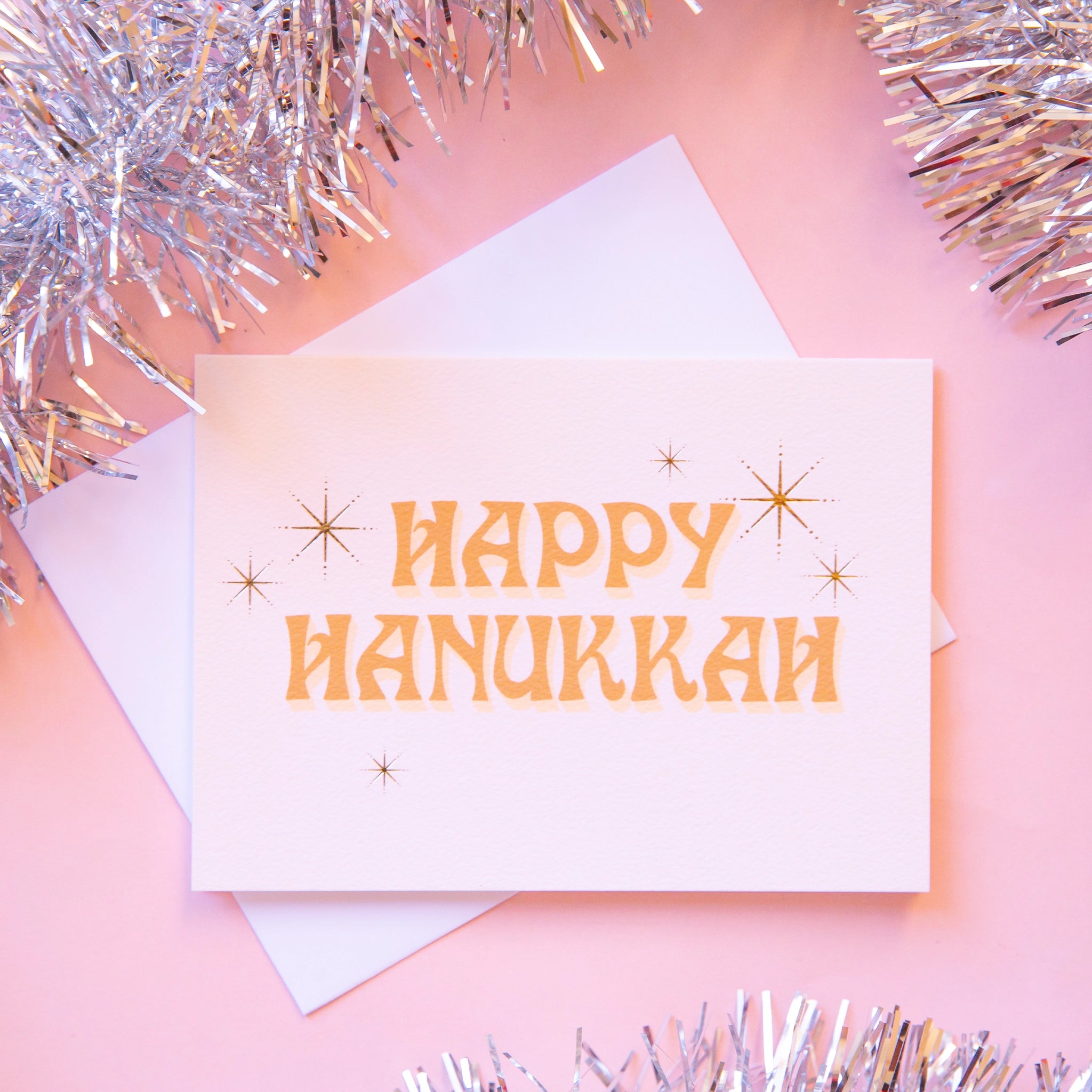 Ivory card that reads &#39;Happy Hanukkah&#39; in retro orange lettering. The text is accented with gold foil stars twinkling around.