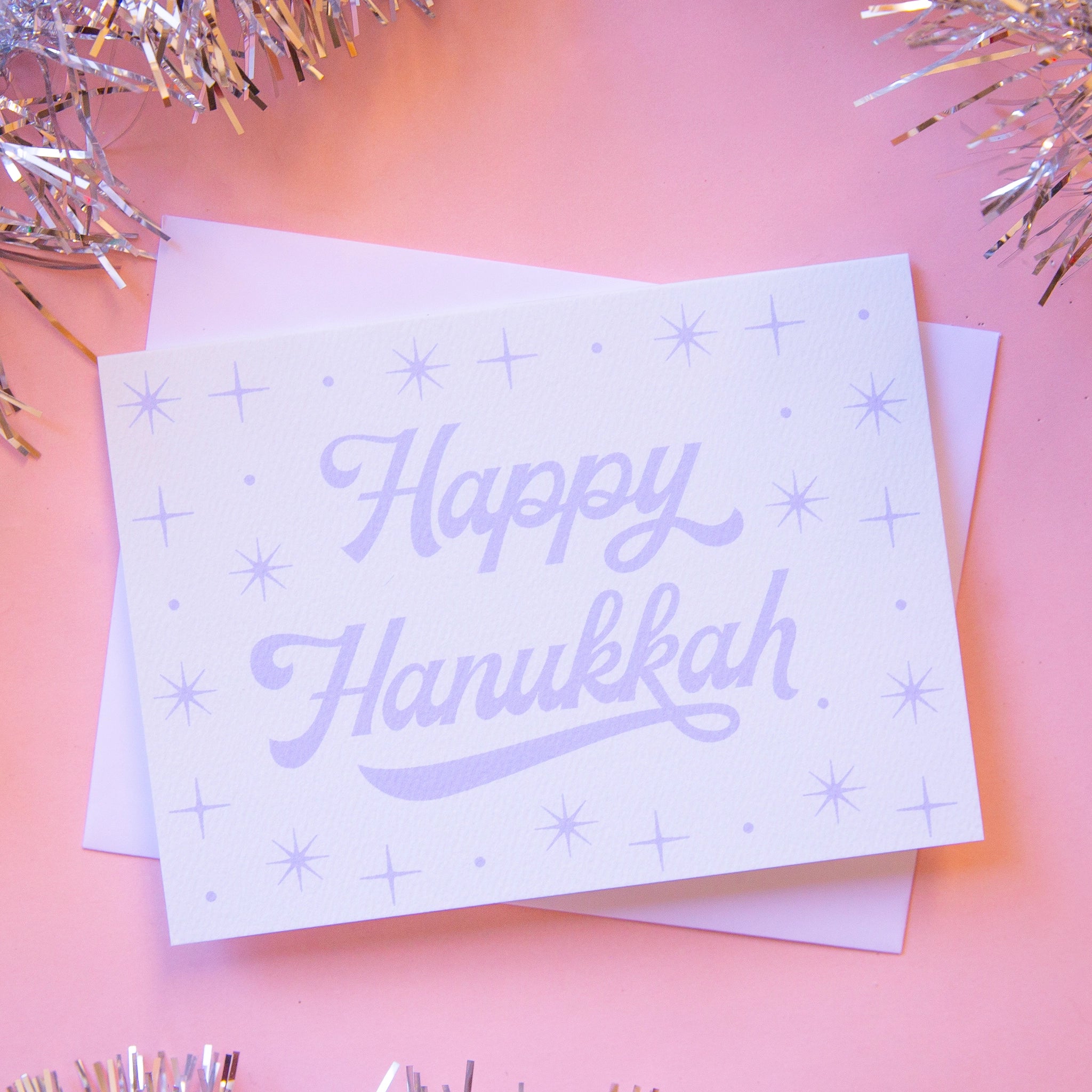 On a pink background is a light blue card that reads, &quot;Happy Hanukkah&quot; with sparkling stars designs around it.