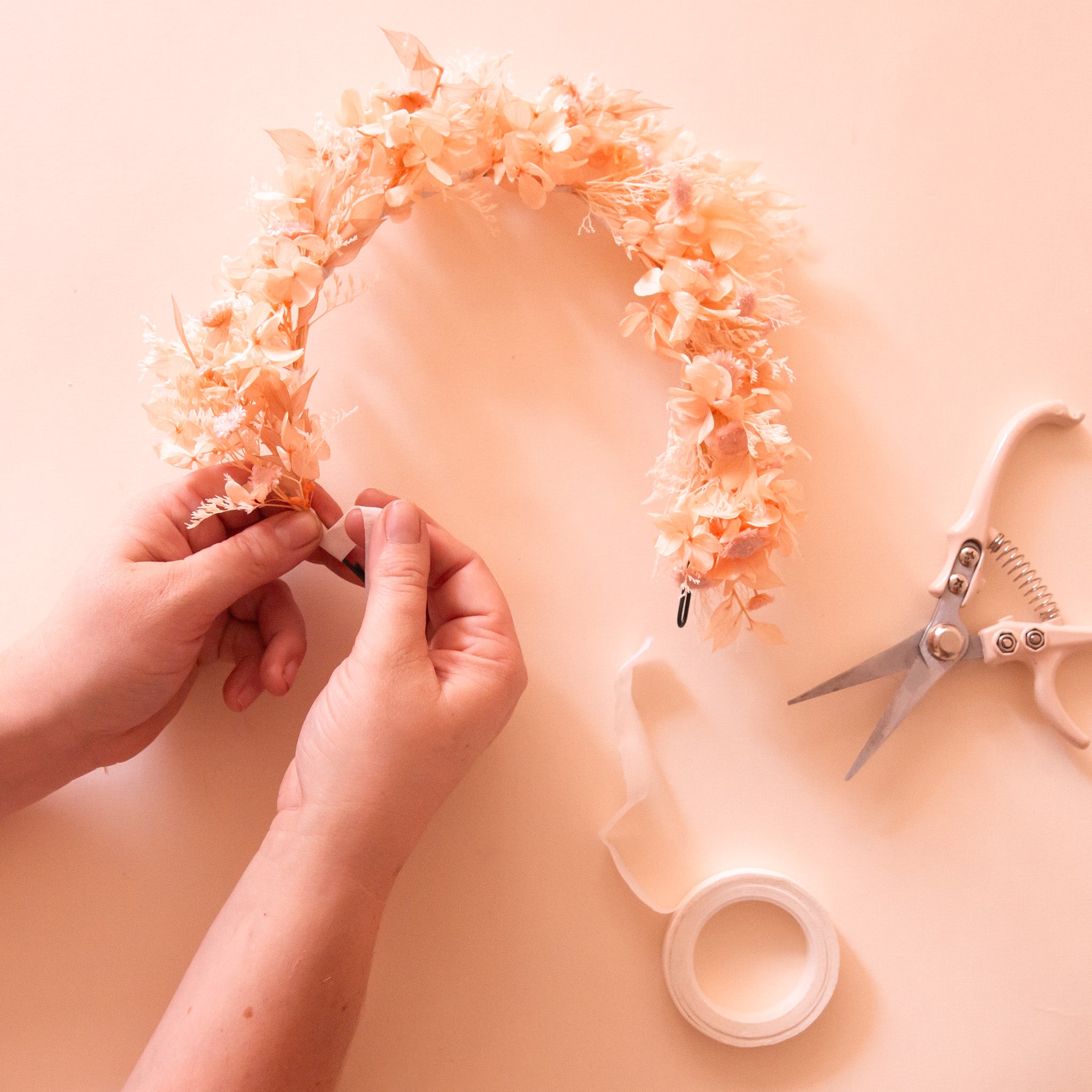 A neutral dried floral flower crown being crafted by someone&#39;s hands next to some ribbon and pruning shears. 