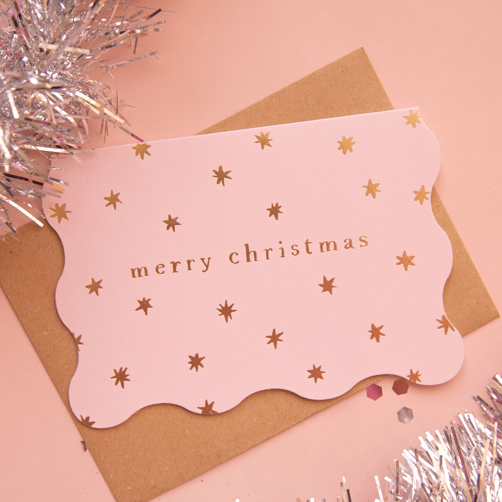 On a pink background is a pink scalloped edge card with gold foiled stars and text that reads, "merry christmas".