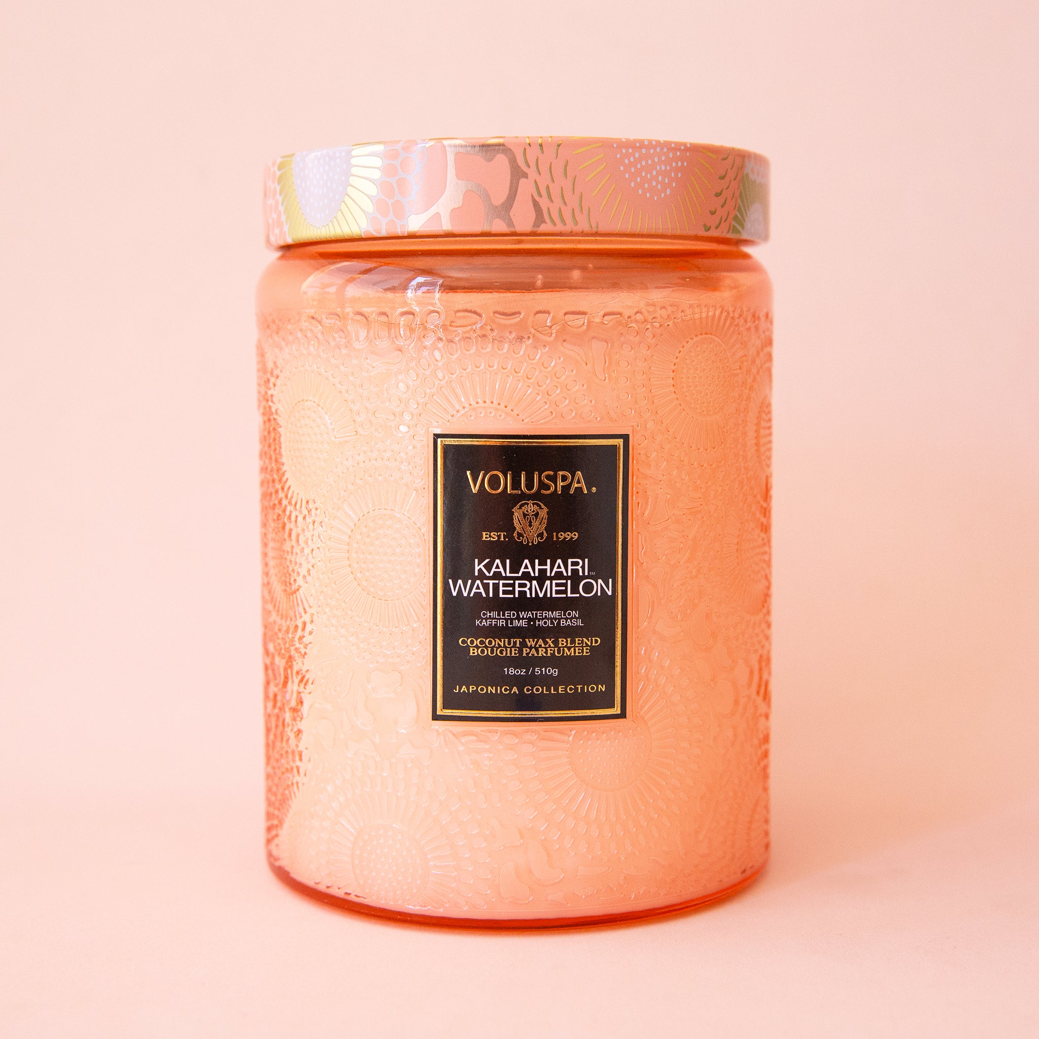 Pink colored embossed candle with patterned colorful lid. The embossed pattern and lid have an abstract floral design with use of the colors peach, gold, and white.