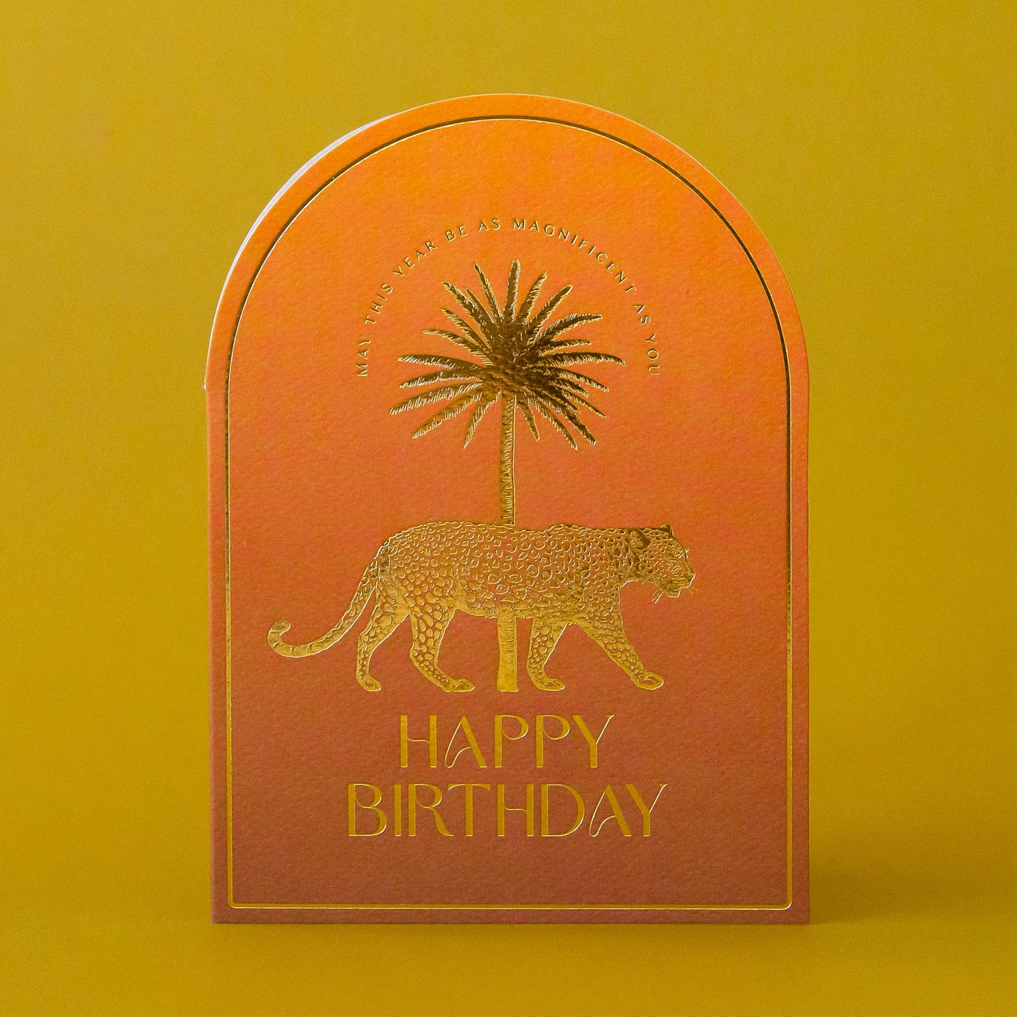 An orange arched card with gold foiled text that reads, &quot;May This Year Be As Magnificent As You Happy Birthday&quot; along with a gold foiled leopard design. 