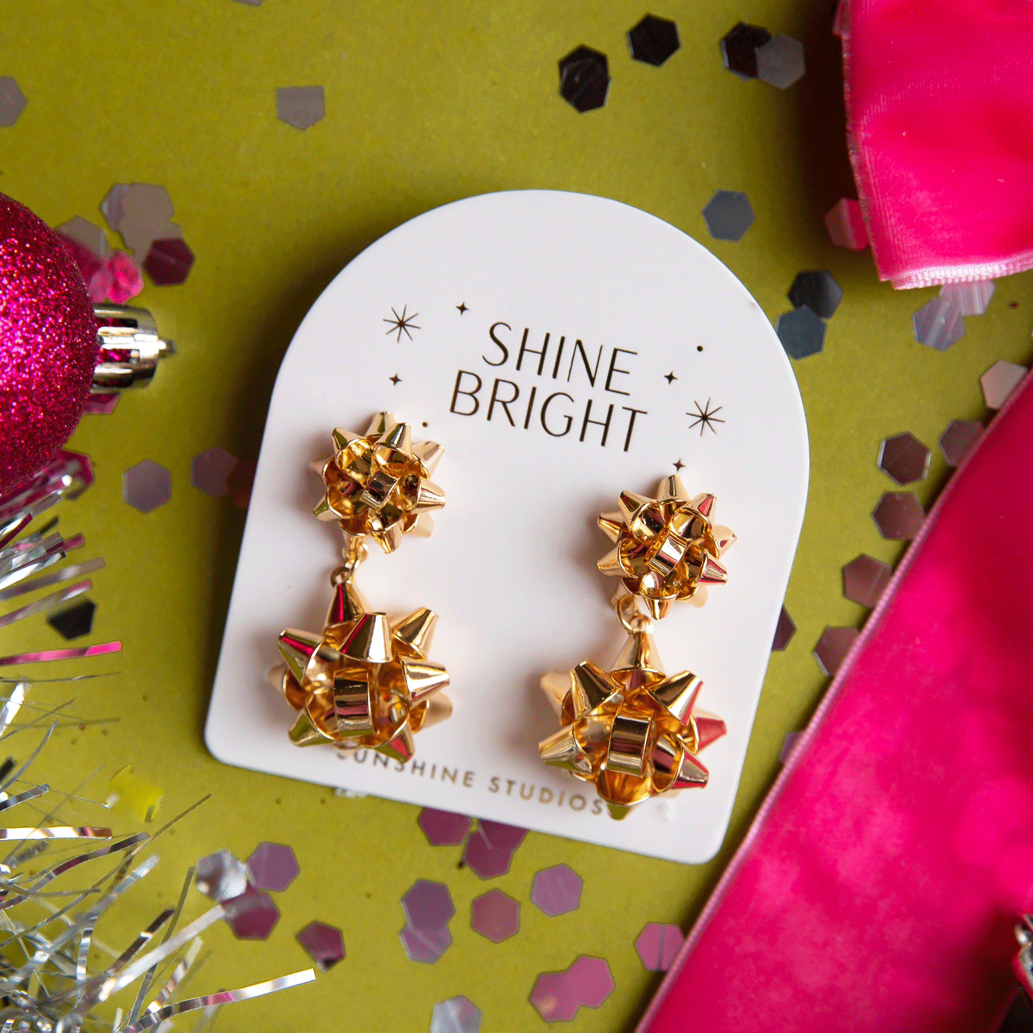 On a green background is a pair of gold holiday bow shaped earrings that have a smaller gold bow and a dangling larger bow attached to the bottom. The packaging reads, &quot;Shine Bright Sunshine Studios&quot;.