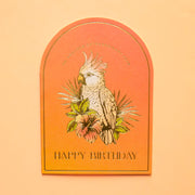 An orange ombre arched shaped card with a bird design on the front and gold foiled text that reads, "To Another Extraordinary Year Happy Birthday".  