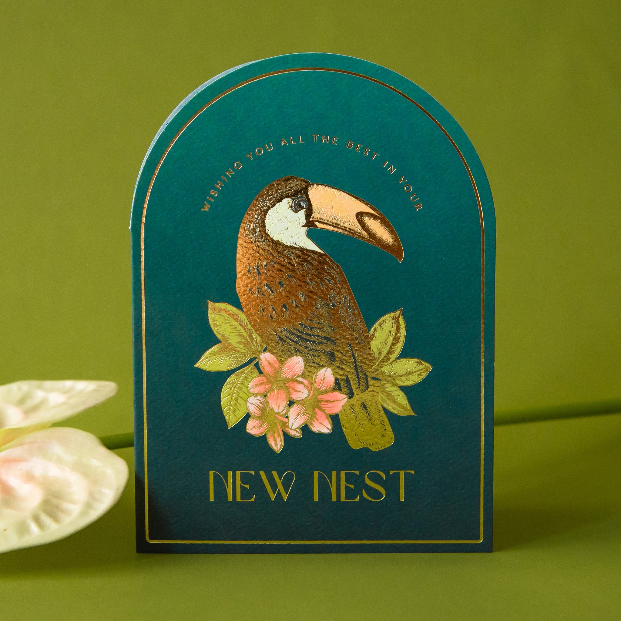 An arched shaped teal blue card, with a gold foiled illustration of a toucan bird and text above and below that reads, "Wishing You All The Best In Your New Nest". 