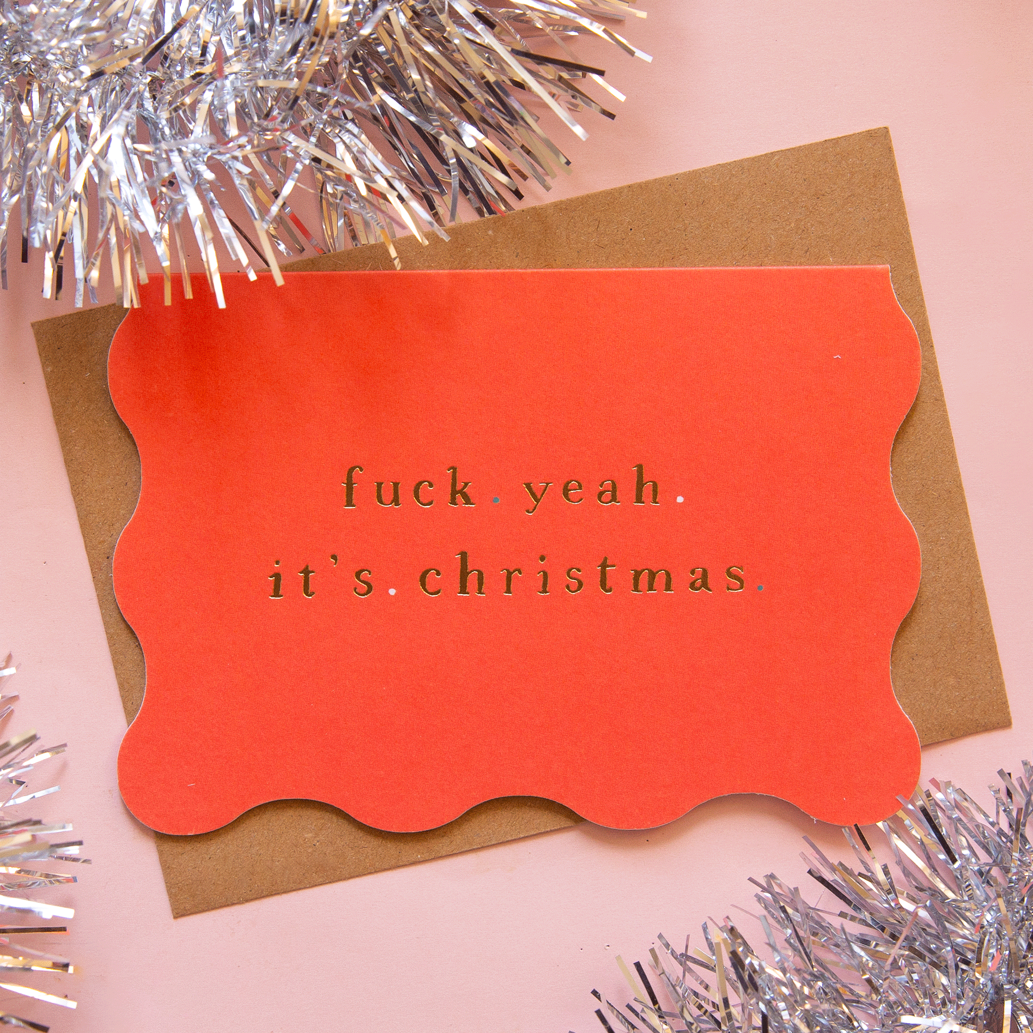 On a pink background is a red scalloped edge card with gold foiled text that reads, "fuck yeah it's christmas".