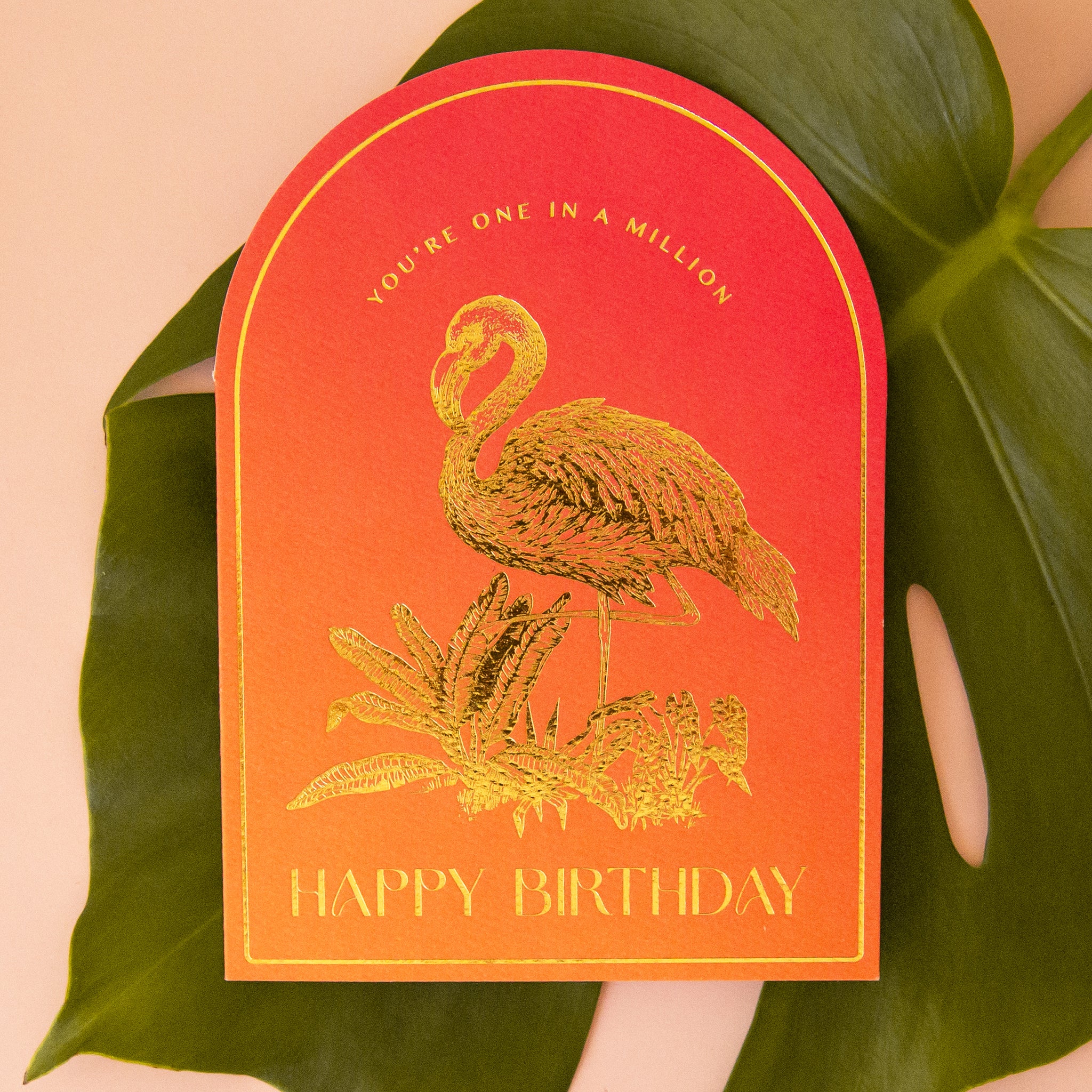 An orange gradient arched shaped card with gold foiled illustrations of a flamingo and text that reads, "You're One In A Million Happy Birthday". 