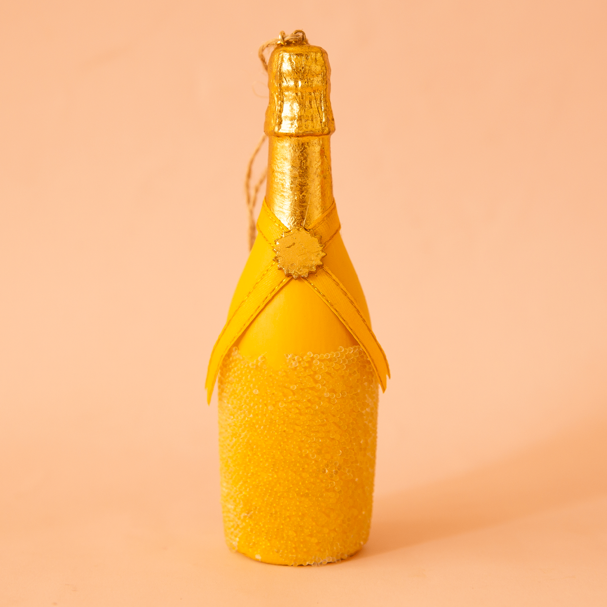 On a pink background is a yellow champagne bottle shaped ornament. 