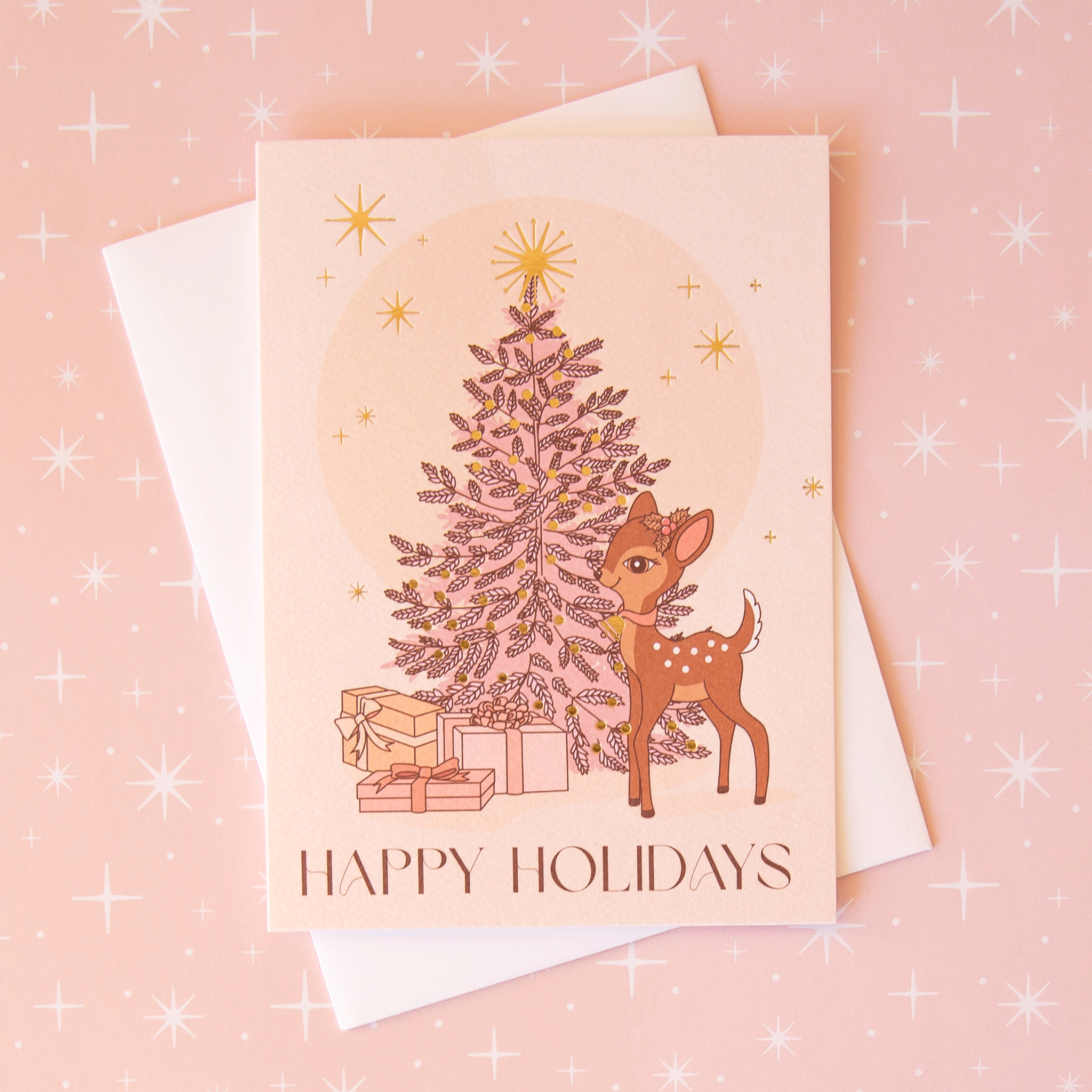 On a pink background is a soft pink greeting card with a Christmas tree and retro deer illustration not he front along with text along the bottom that reads, "Happy Holidays".