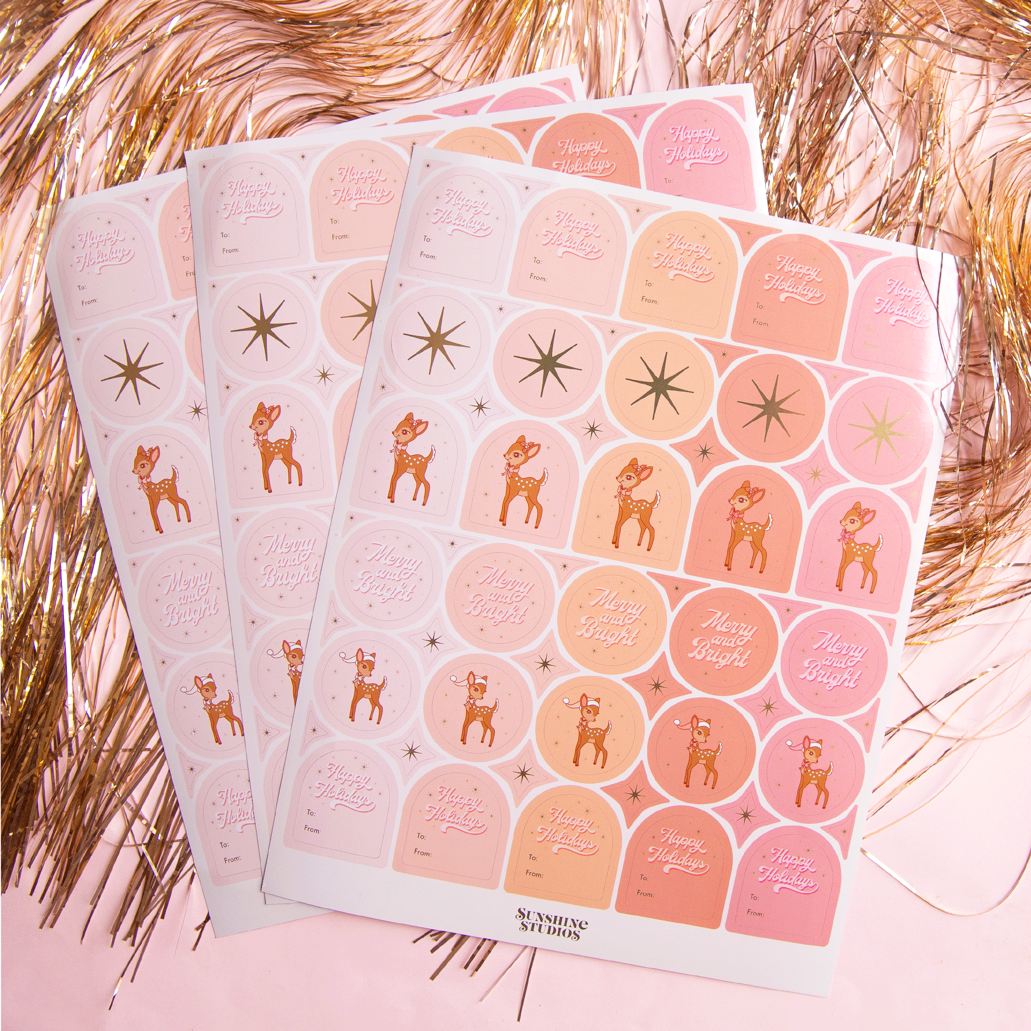 On a pink background is three sheets of gift label stickers with shapes of circles and arches along with text that reads, &quot;Happy Holidays&quot;, a gold star, or a retro deer graphic.