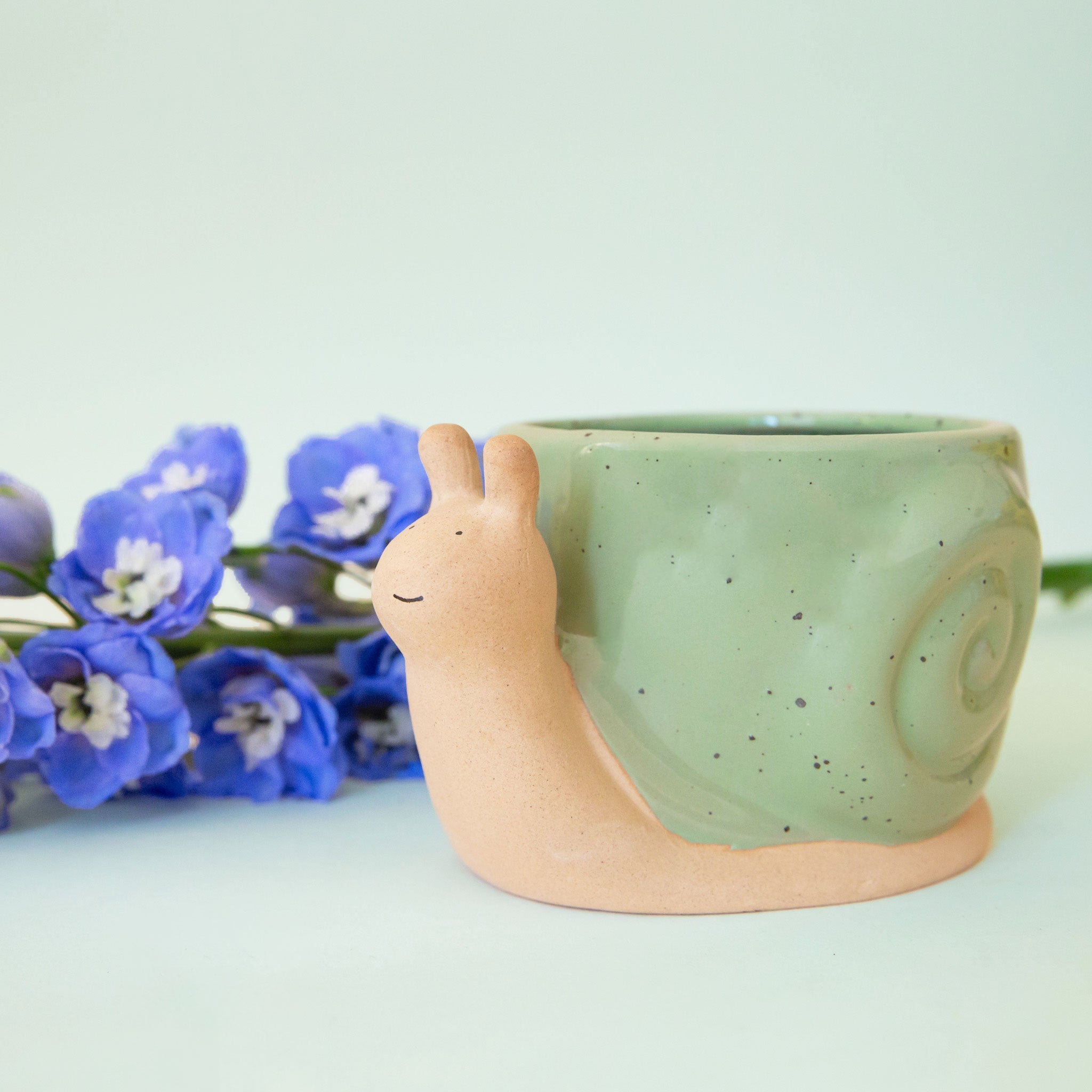 On a tan background is a ceramic planter in the shape of a snail with smiling face and a swirly green "shell".