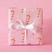 A single sheet of gift wrap with a blush background and a repeating red and white candy cane print. The gift wrap is reversible and the other side features a light purple / lavender background with an all over red and white candy cane design but the candy canes are slightly smaller than the reverse. Present is wrapped with a silver tinsel ribbon and surrounded by snow.