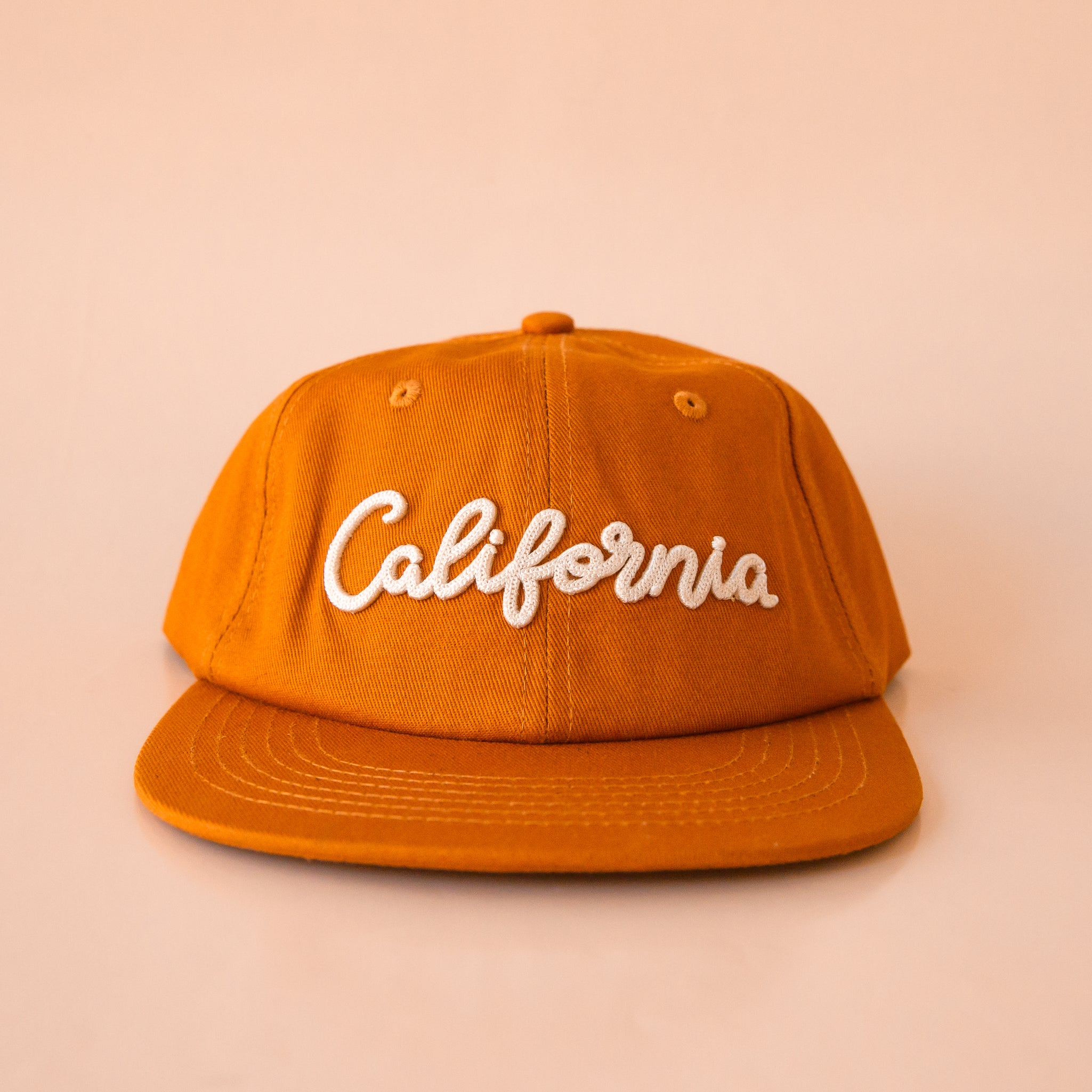 A burnt orange flat brimmed baseball hat with ivory embroidered text that reads, "California" across the front.  