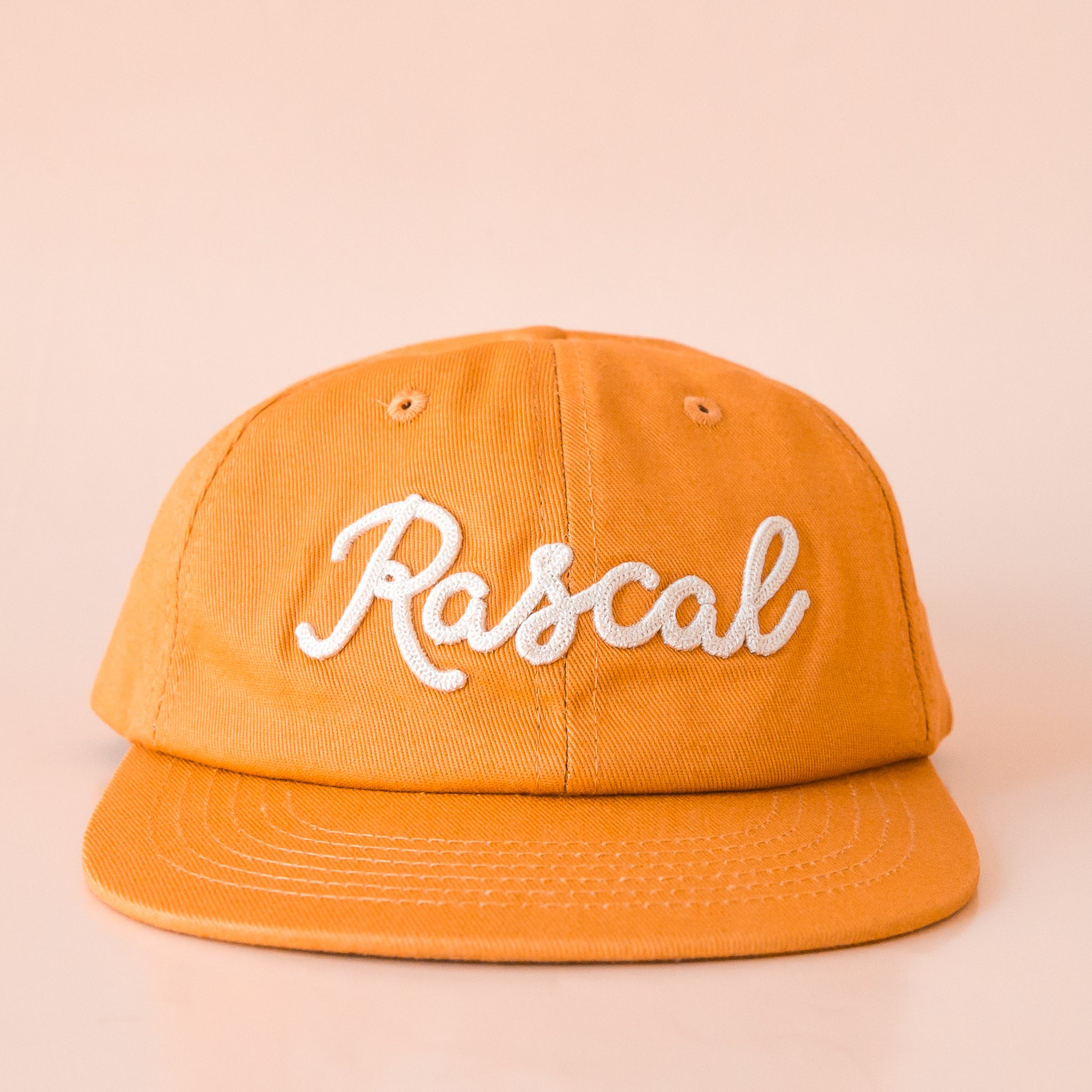 A burnt orange flat brimmed hat with white embroidered text that reads, &quot;Rascal&quot;.