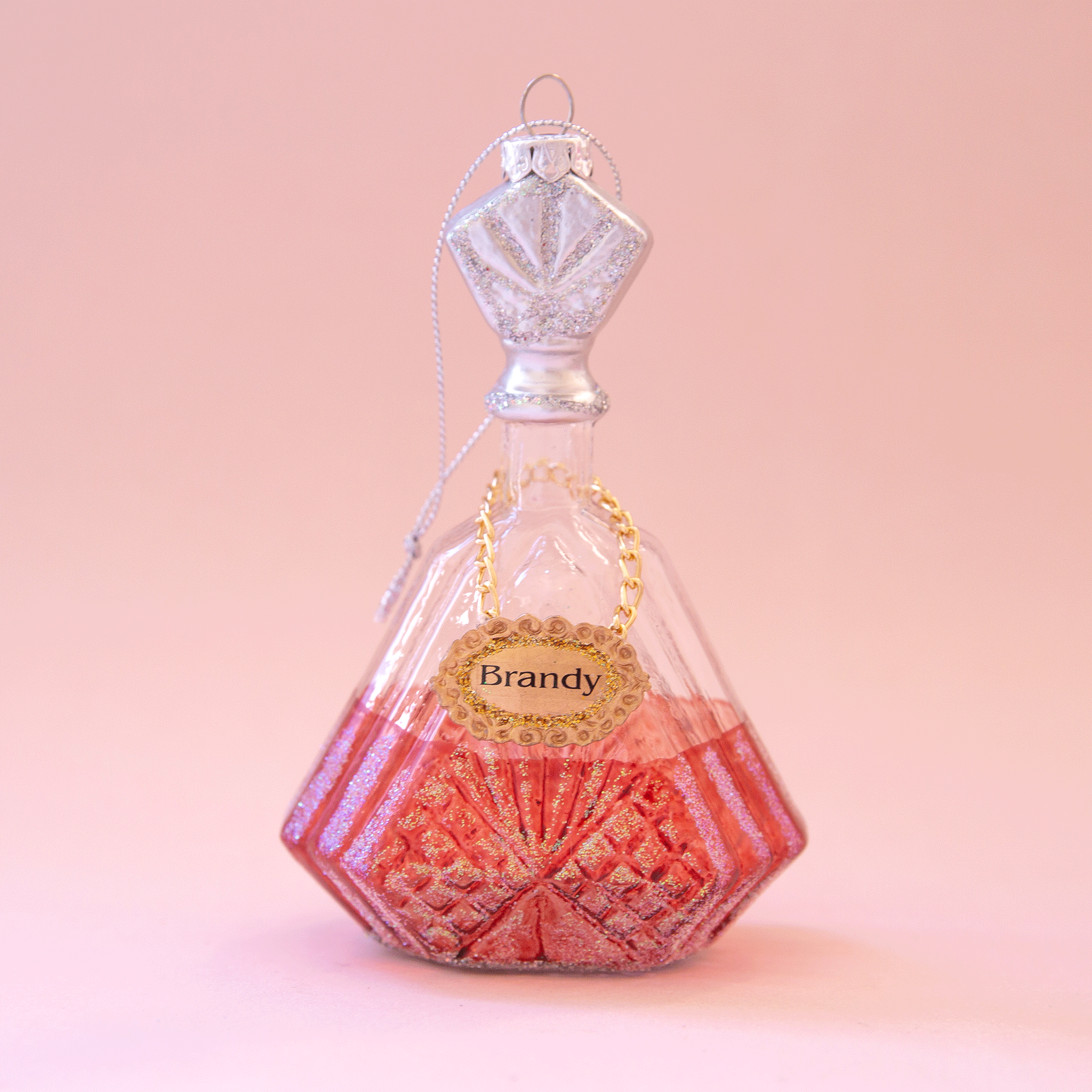 A glass decanter ornament with a gold label that reads, "Brandy" along with a pinkish base. The stopper to the decanter is silver and detailed with glitter.