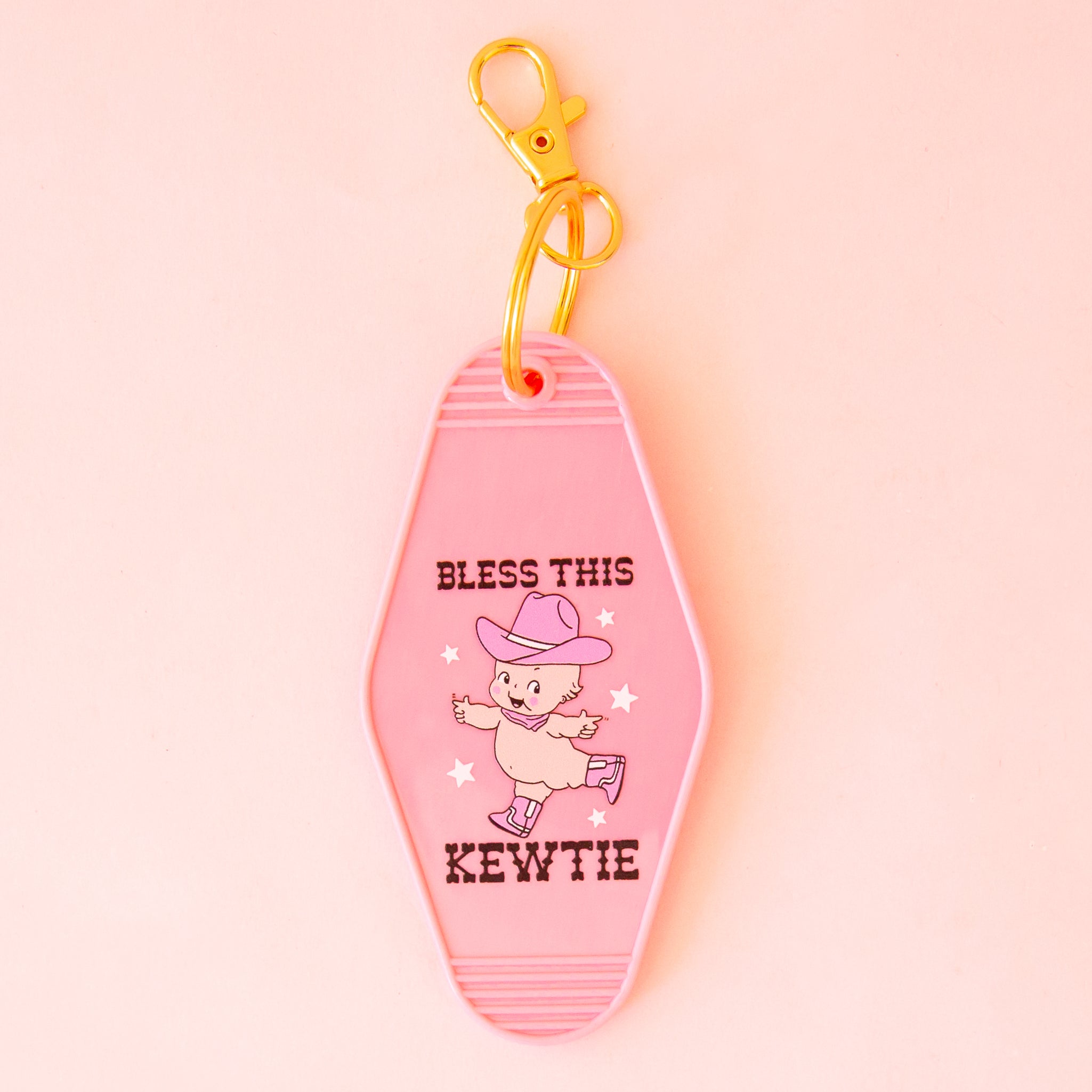A pink motel style keychain with a kewtie baby design wearing pink cowgirl hat and boots with text that reads, "Bless This Kewtie". 