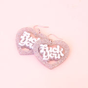 A pair of silver glitter heart shaped earrings with white text in the center that reads, "Fuck You".