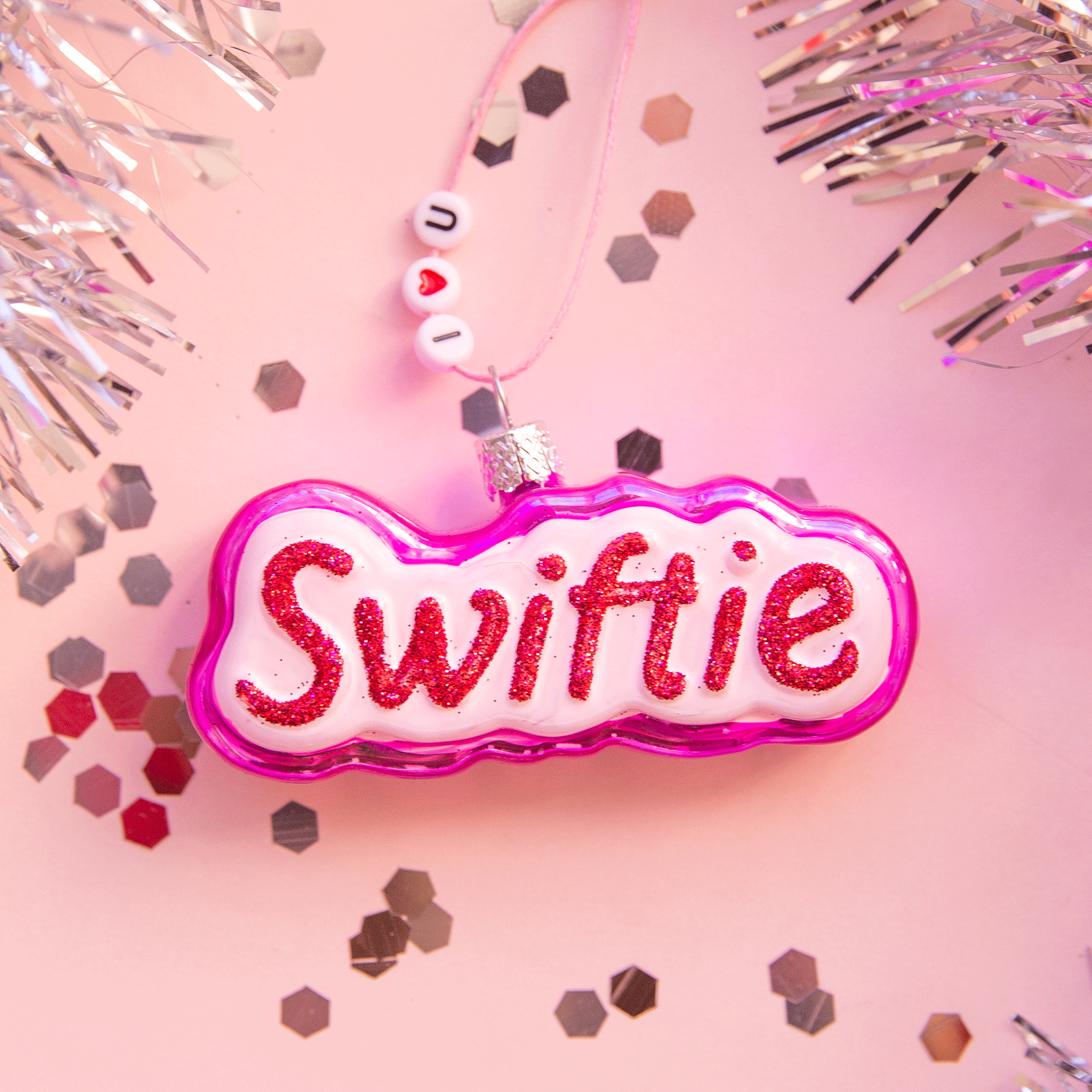 pink ornament that reads "swiftie" in red glitter writing. background is pink with silver tinsel and confetti.