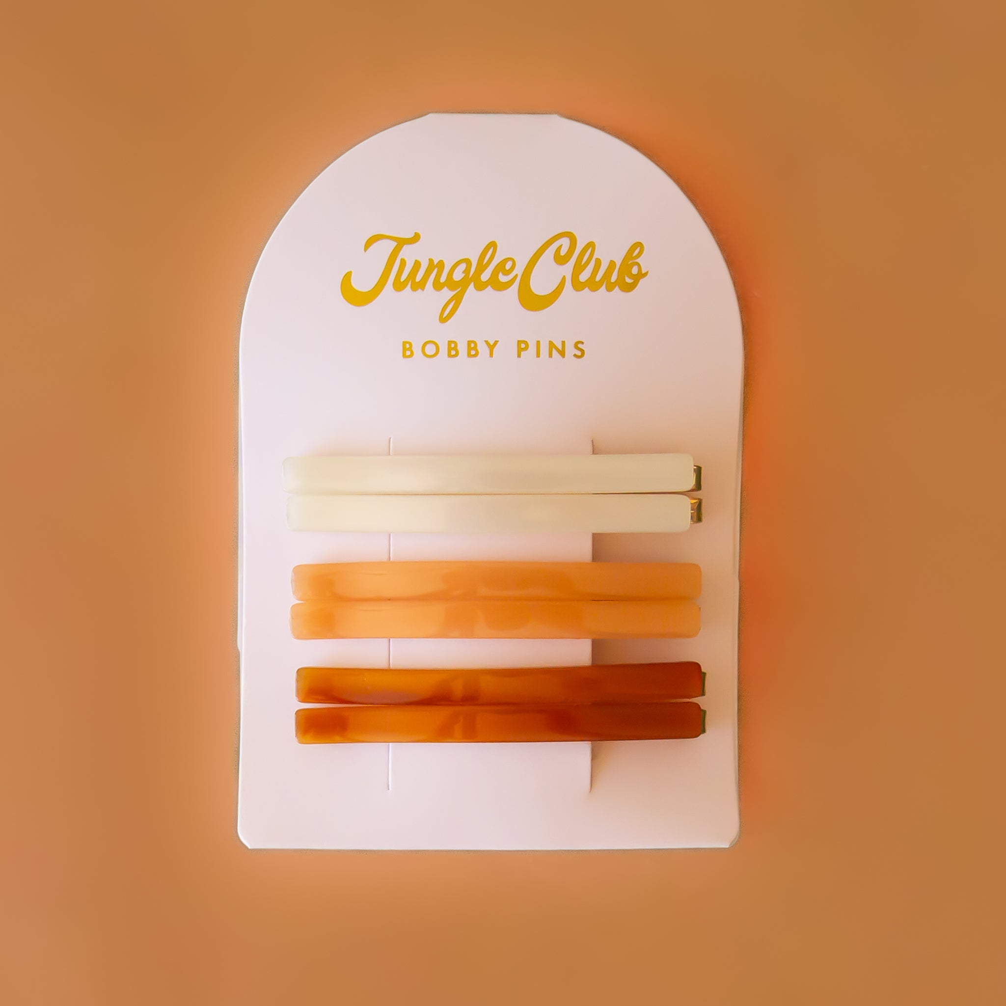 On a brown background is an arched packaging with embossed text at the top that reads, "Jungle Club Bobby Pins" along with six thin hair clips in three colors from an ivory shade to orange to a honey rust color.