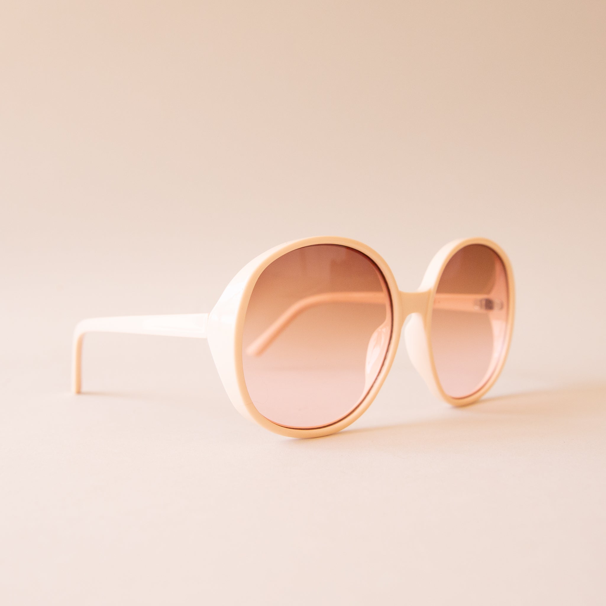 A creamy white pair of round oversized sunglasses with a light brown pinkish lens. 