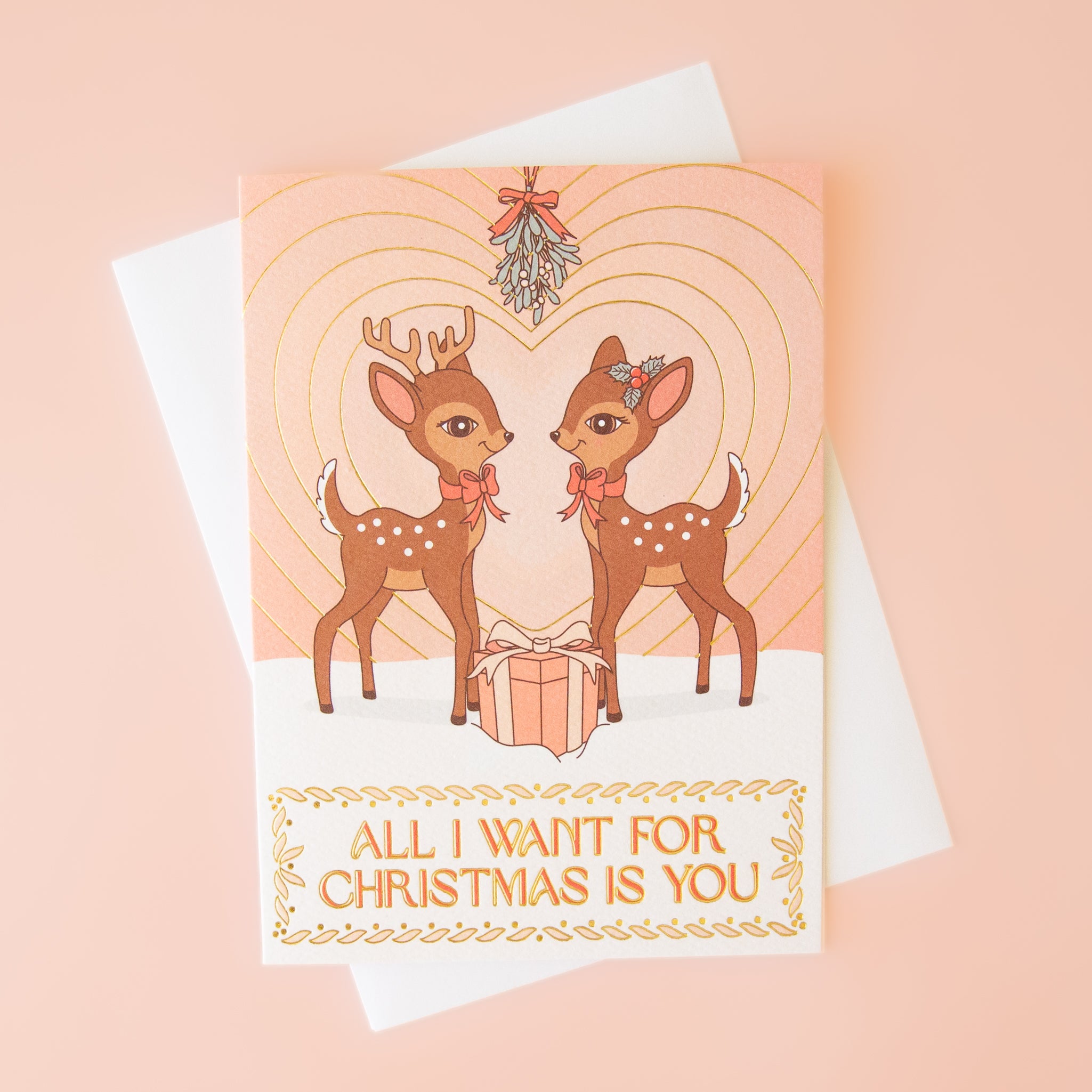 On a neutral background is a light pink card with two brown retro deer looking towards one another underneath mistletoe with a gradient heart in the background and text below the image that reads, &quot;All I Want For Christmas Is You&quot;. 