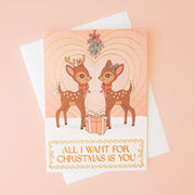 On a neutral background is a light pink card with two brown retro deer looking towards one another underneath mistletoe with a gradient heart in the background and text below the image that reads, "All I Want For Christmas Is You". 