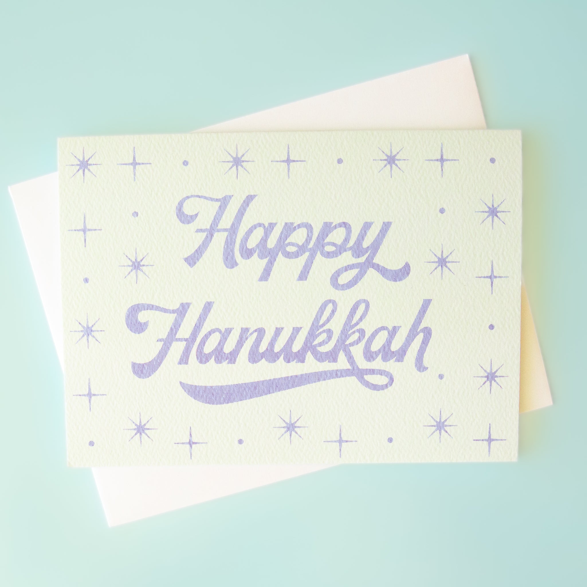 A light blue card that reads, &quot;Happy Hanukkah&quot; with sparkling stars designs around it. 