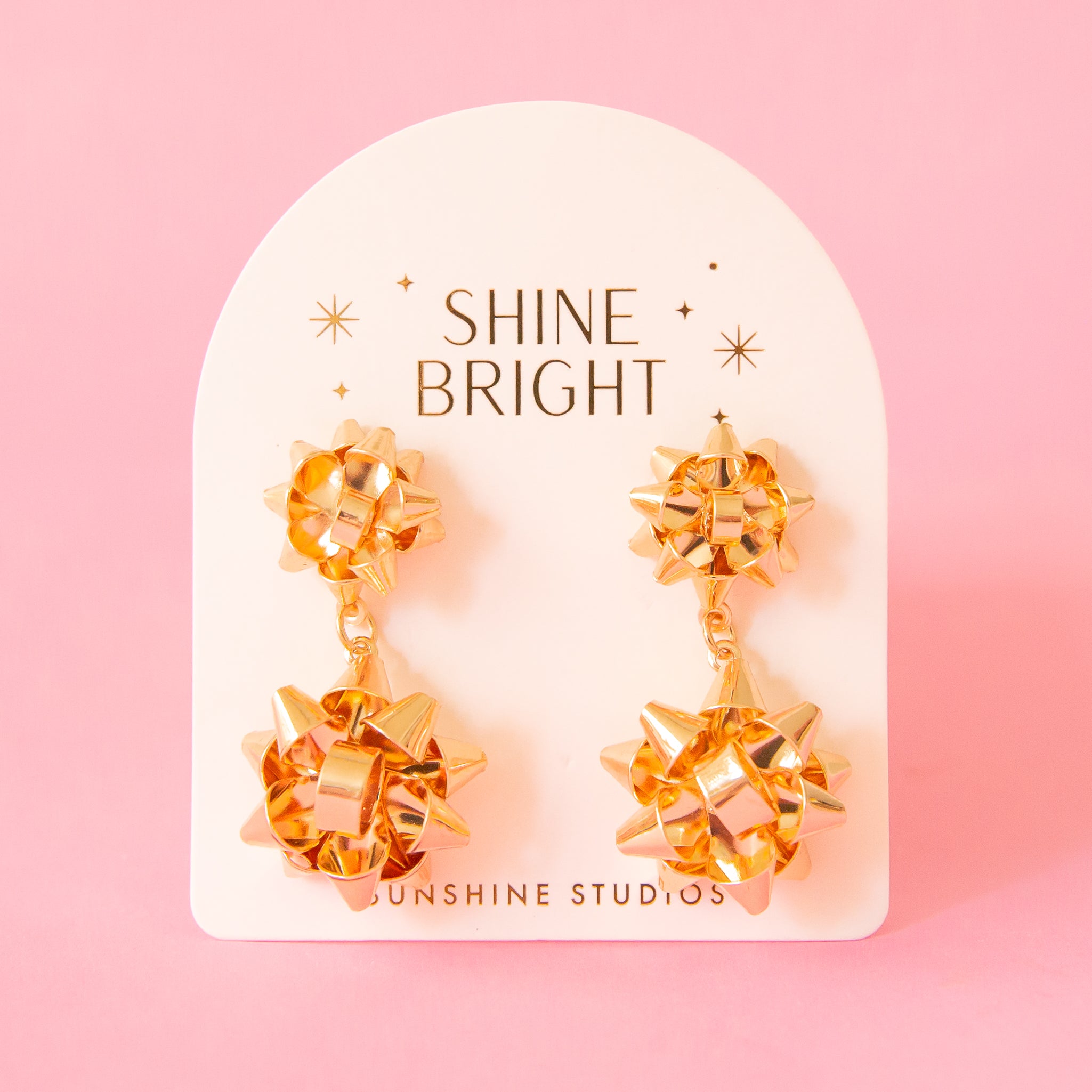 A pair of gold holiday bow shaped earrings that have a smaller gold bow and a dangling larger bow attached to the bottom. The packaging reads, &quot;Shine Bright Sunshine Studios&quot;.