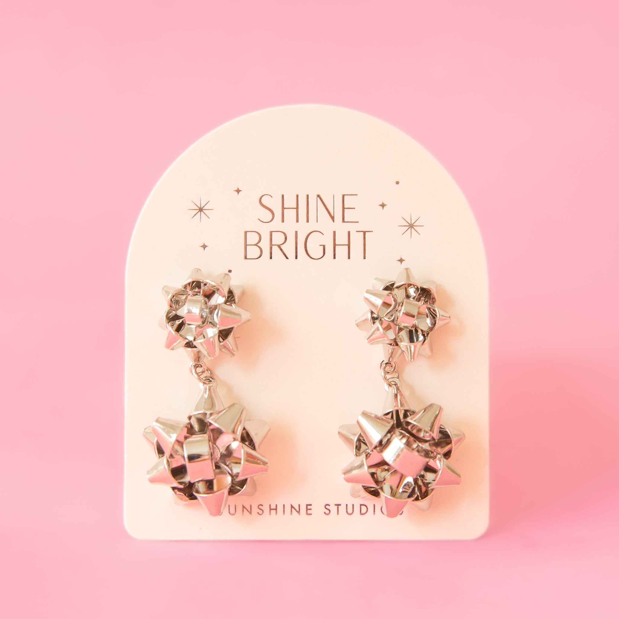 A pair of silver holiday bow shaped earrings that have a smaller gold bow and a dangling larger bow attached to the bottom. The packaging reads, &quot;Shine Bright Sunshine Studios&quot;.