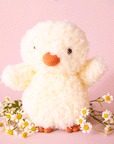 On a pink background is a super light yellow fuzzy chick stuffed animal toy with an orange beak and feet.