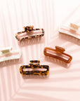 On a light pink background is a variety of rectangle claw clips in tortoise shades, an amber color and two different whites.