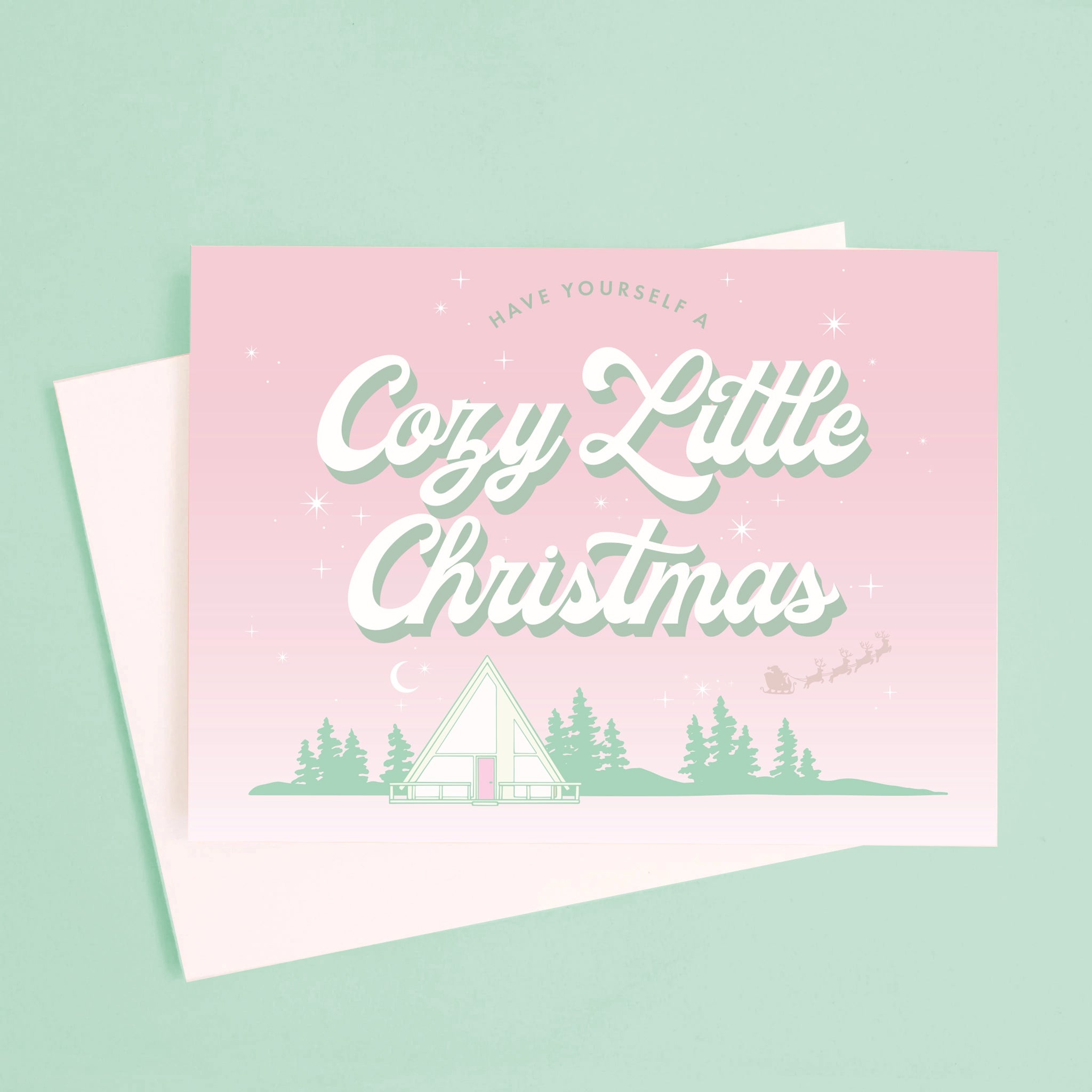 A pink and mint green Christmas card with an A-frame graphic and text above that reads, &quot;Have Yourself A Cozy Little Christmas&quot;.