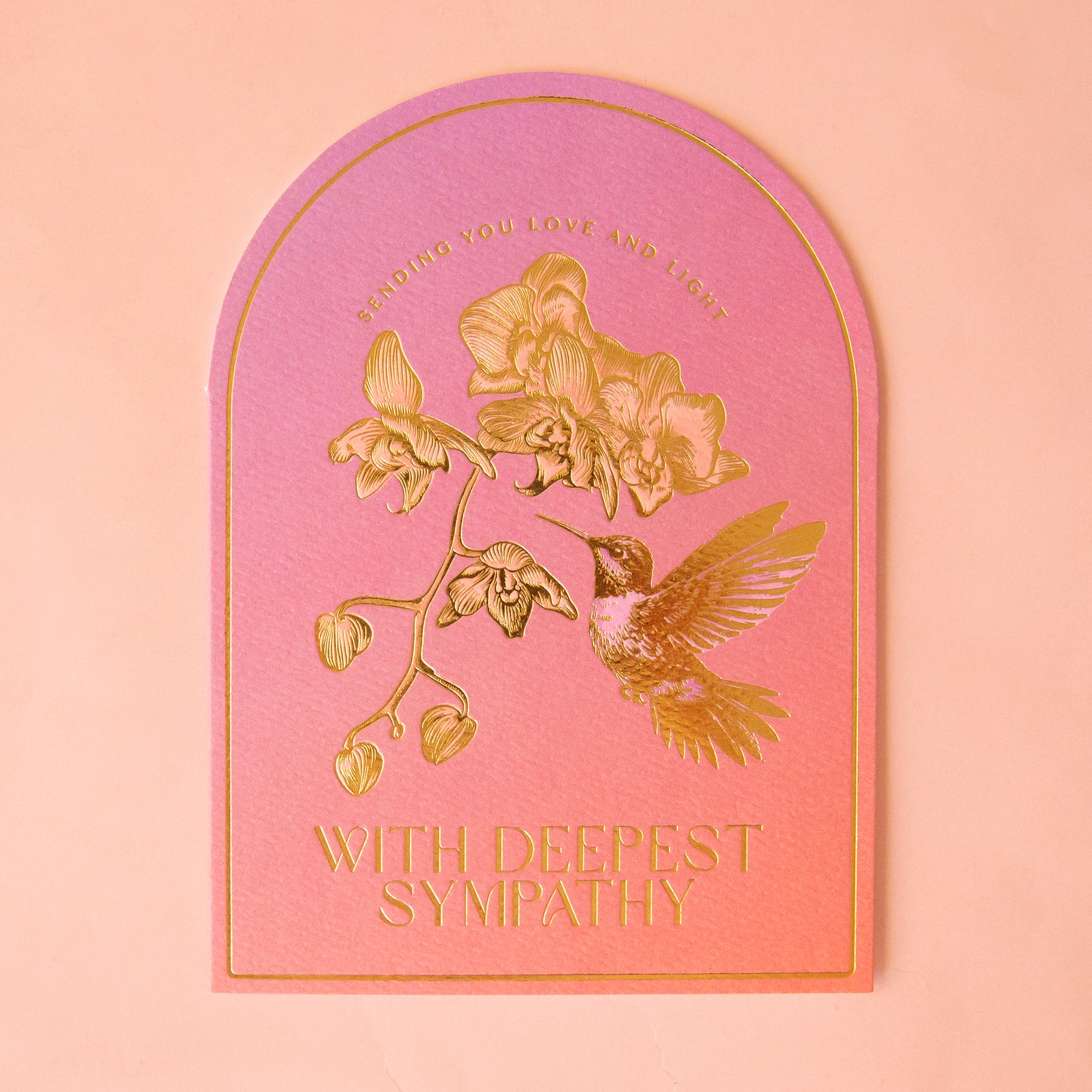 On a peach background is a pink and orange ombre card with gold foiled text that reads, &quot;Sending You Love And Light With Deepest Sympathies&quot; along with a design of a hummingbird.  