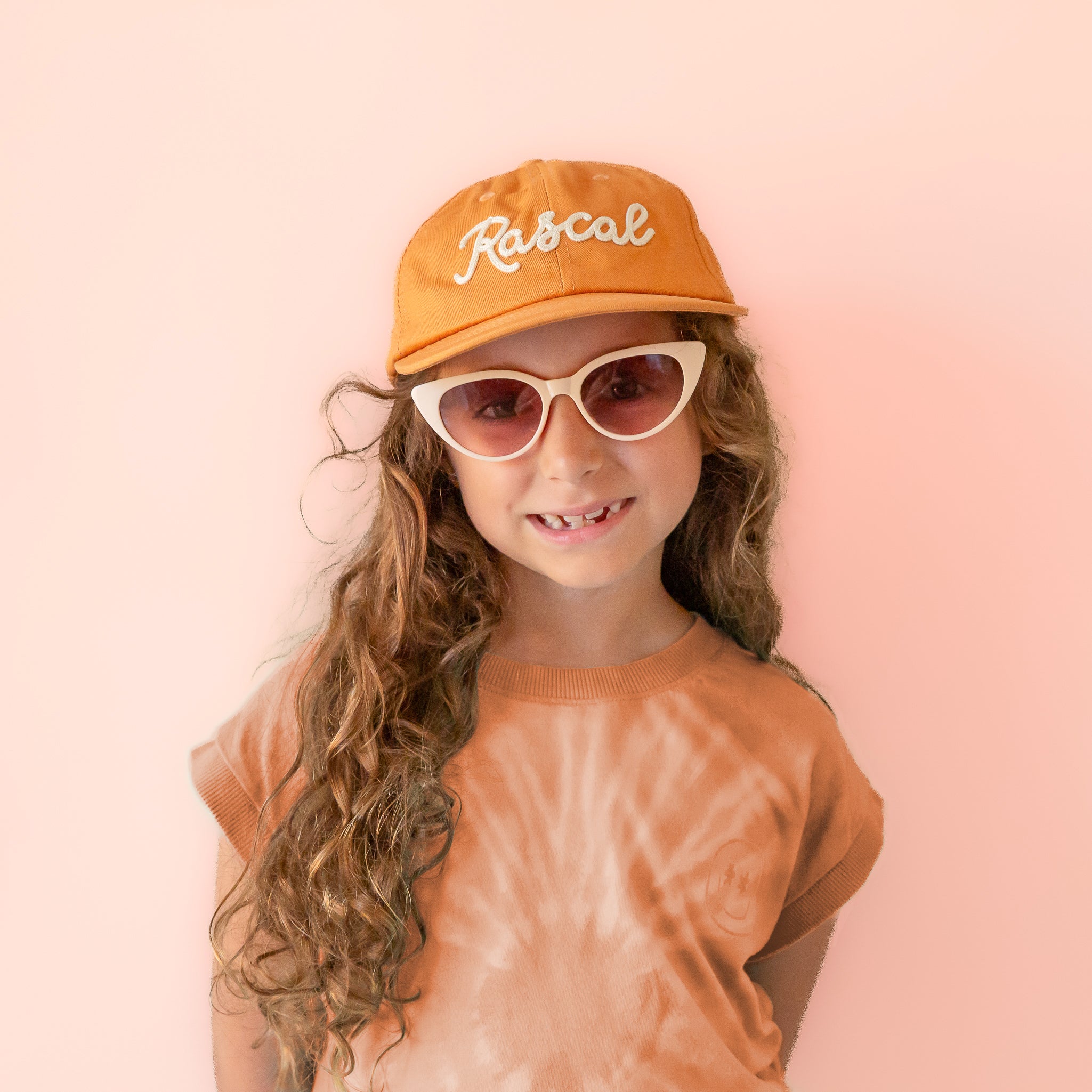 A burnt orange flat brimmed hat with white embroidered text that reads, &quot;Rascal&quot;.