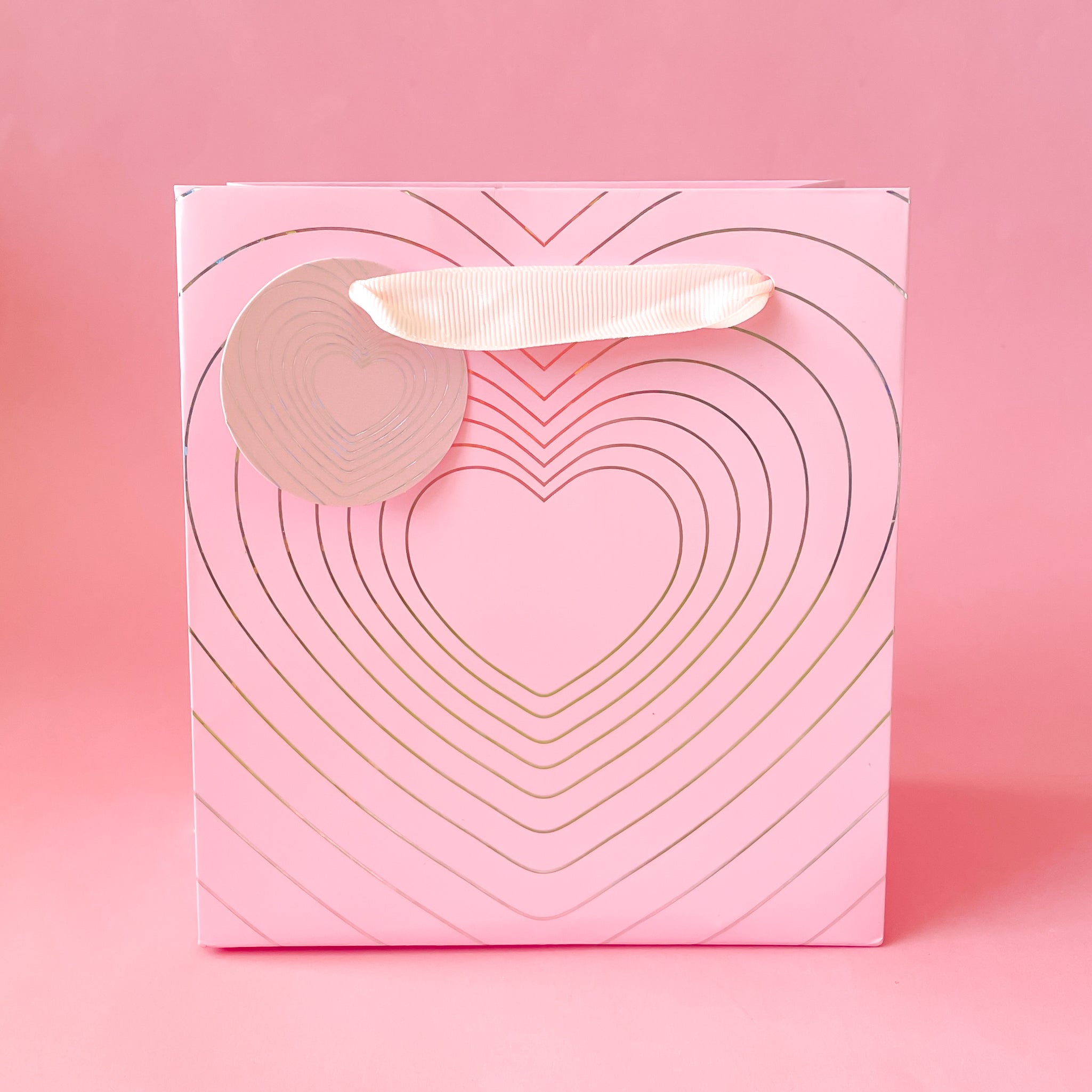 On a light pink background is a light pink bag with a silver radiating heart design and ribbon handles.