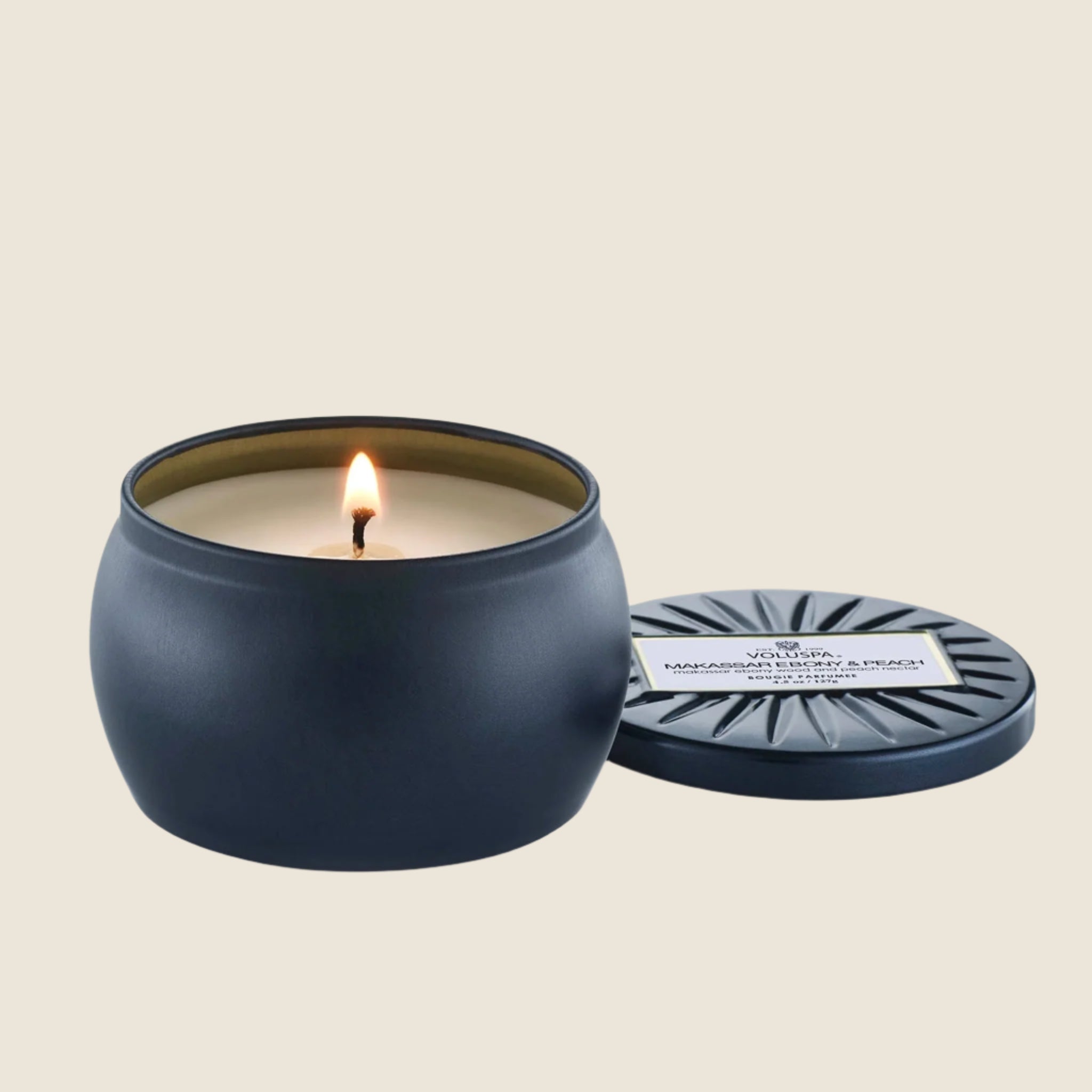 On a cream background is a round tin single wick candle in a navy blue shade along with a label on the lid that reads, "Voluspa Makassar Ebony & Peach".