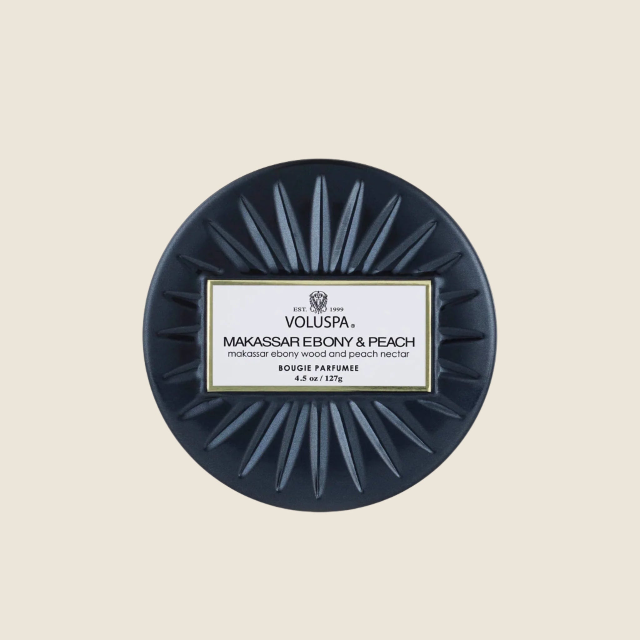 On a cream background is a round tin single wick candle in a navy blue shade along with a label on the lid that reads, "Voluspa Makassar Ebony & Peach".