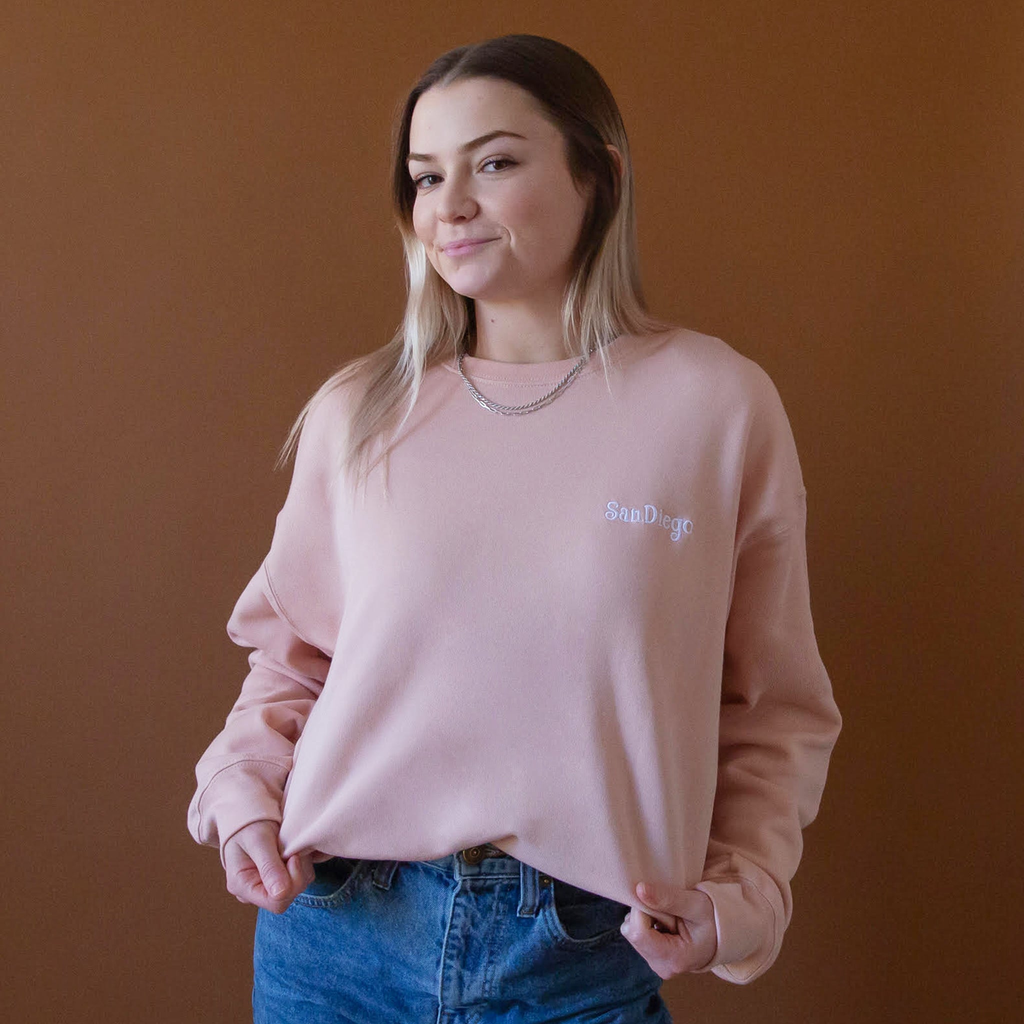 On a peach background is a peachy pink pull over sweatshirt with a white embroidered "San Diego" on the front.