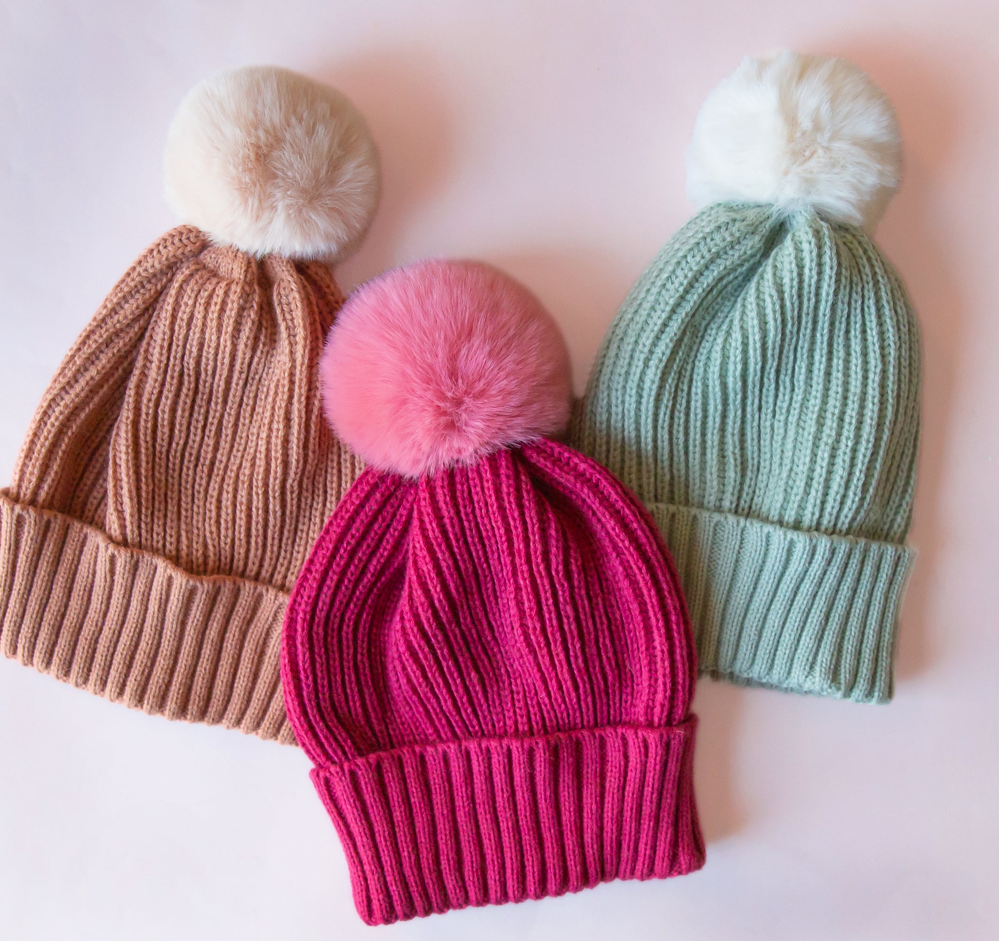 Three different colored beanies with poms at the top. The colors are a tan, a hot pink and a mint green. 